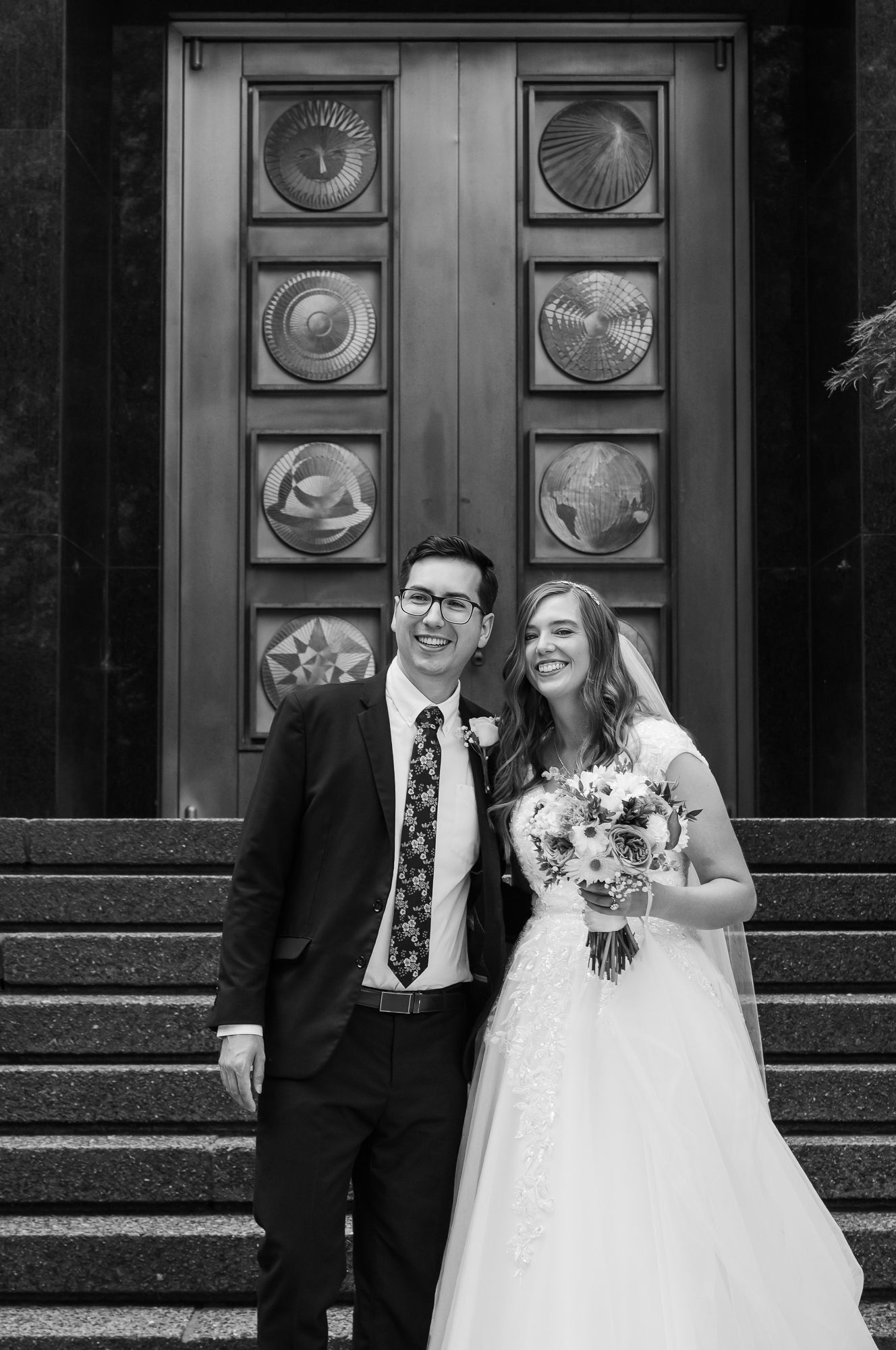 Black and white photo of a smiling bride and groom posing on church steps with ornate doors behind them.