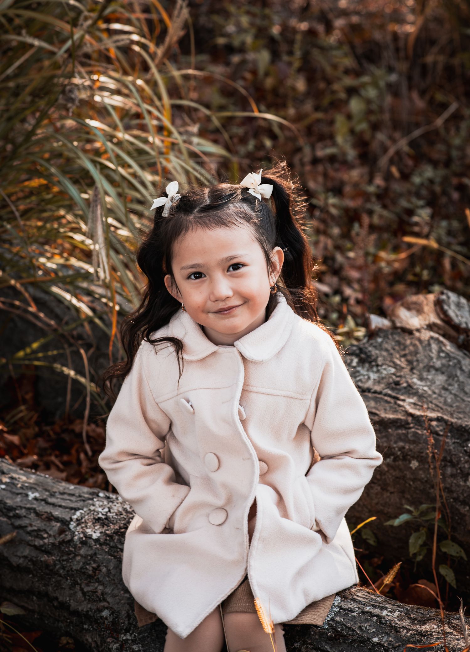A young girl in a cream coat with white bows sits on a log surrounded by autumn foliage.