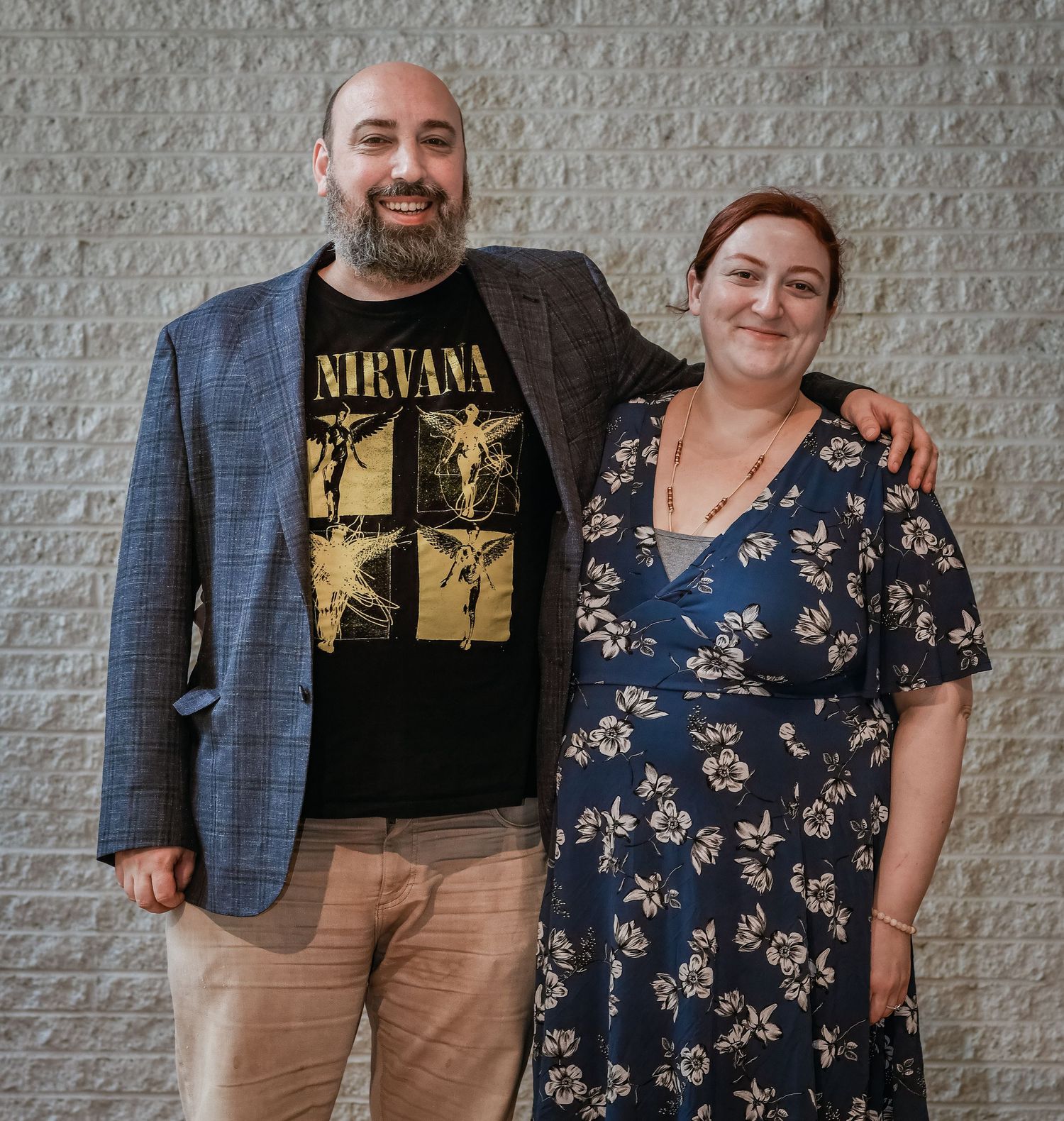 A couple poses against a grey wall, with one wearing a Nirvana t-shirt and blazer.