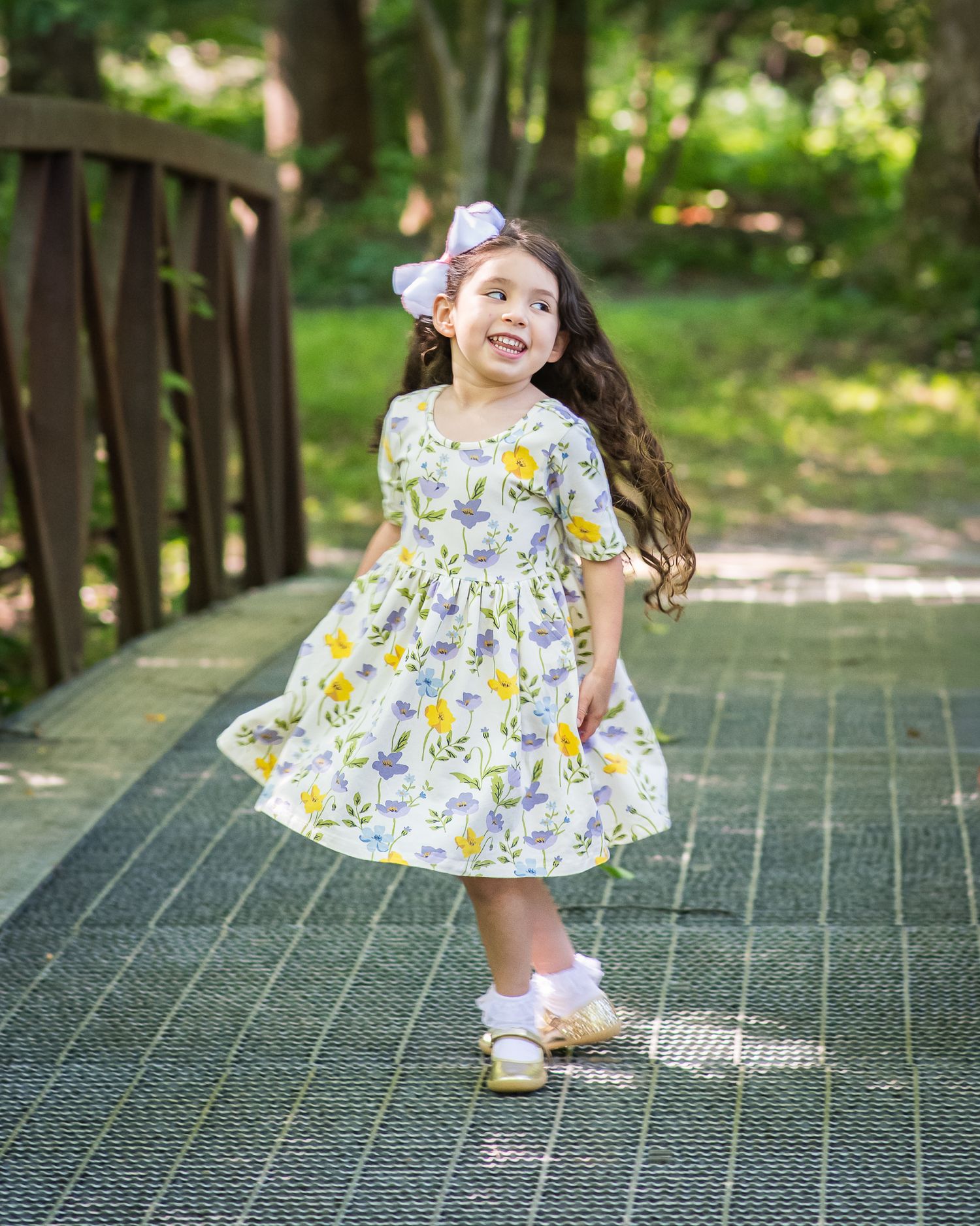 Young girl in floral dress with bow twirling on garden bridge, smiling joyfully outdoors.
