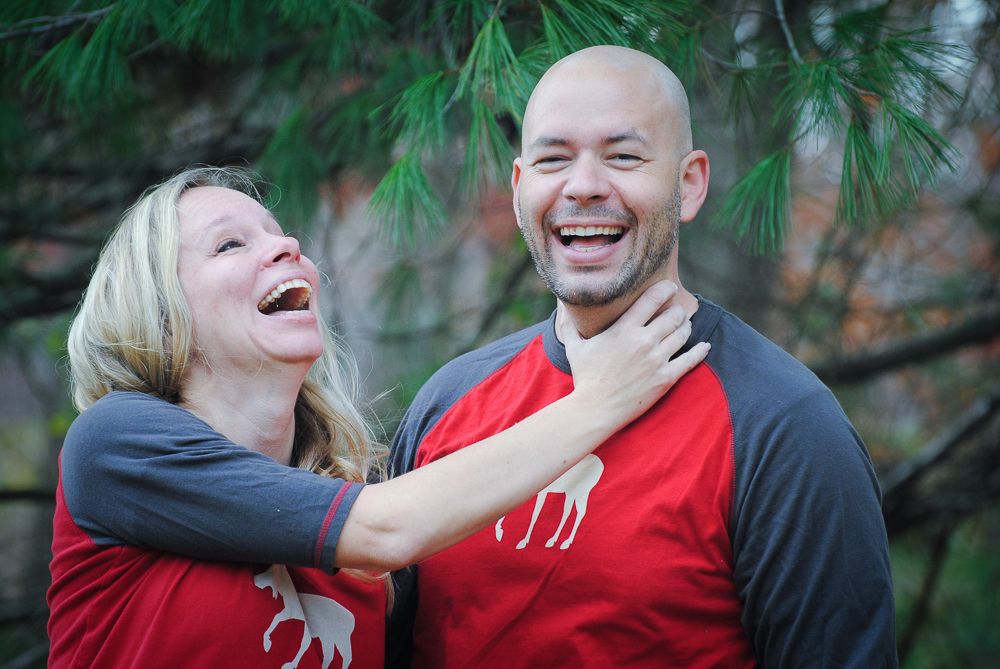 A laughing couple in matching red and gray moose shirts share a joyful moment outdoors among pine trees.
