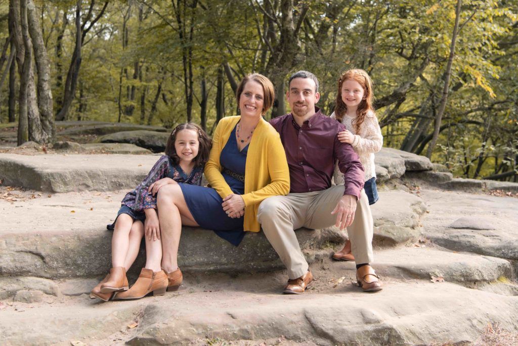 A family of four sits together on a rocky outcrop in a wooded area during autumn.