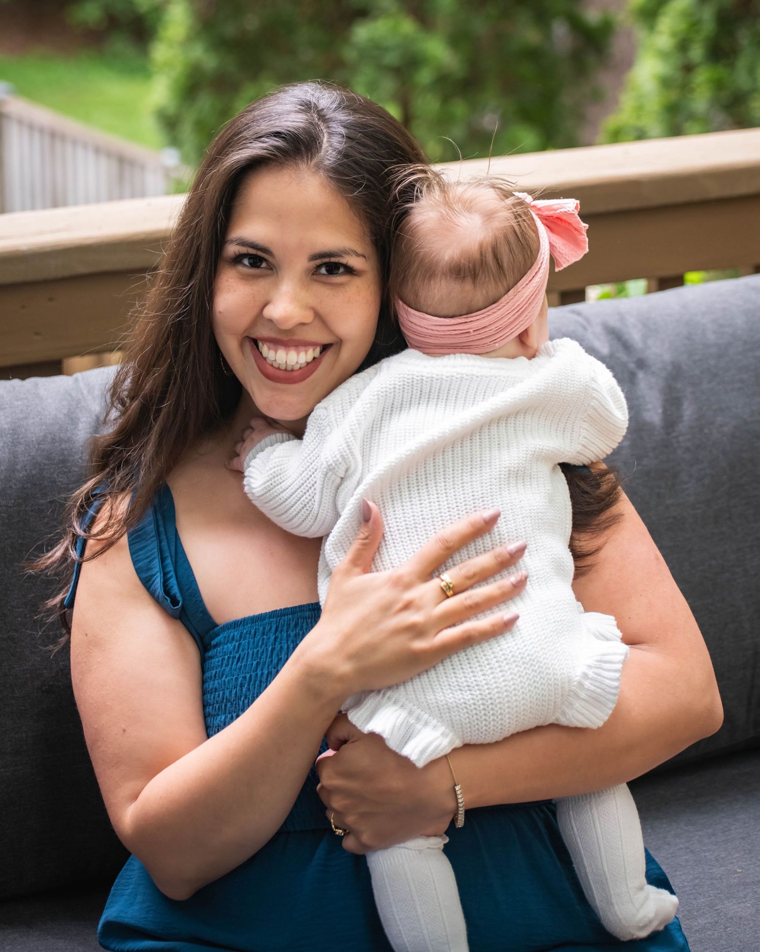 Smiling woman in teal dress holds baby girl in white knit outfit with pink bow on outdoor patio.