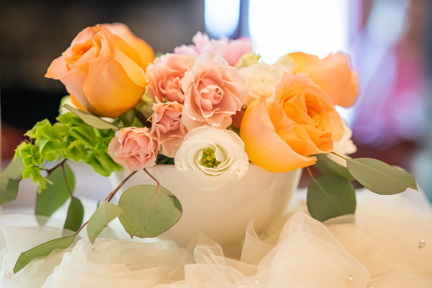 White vase with peach roses, pink carnations, white ranunculus, and eucalyptus leaves as a floral centerpiece.