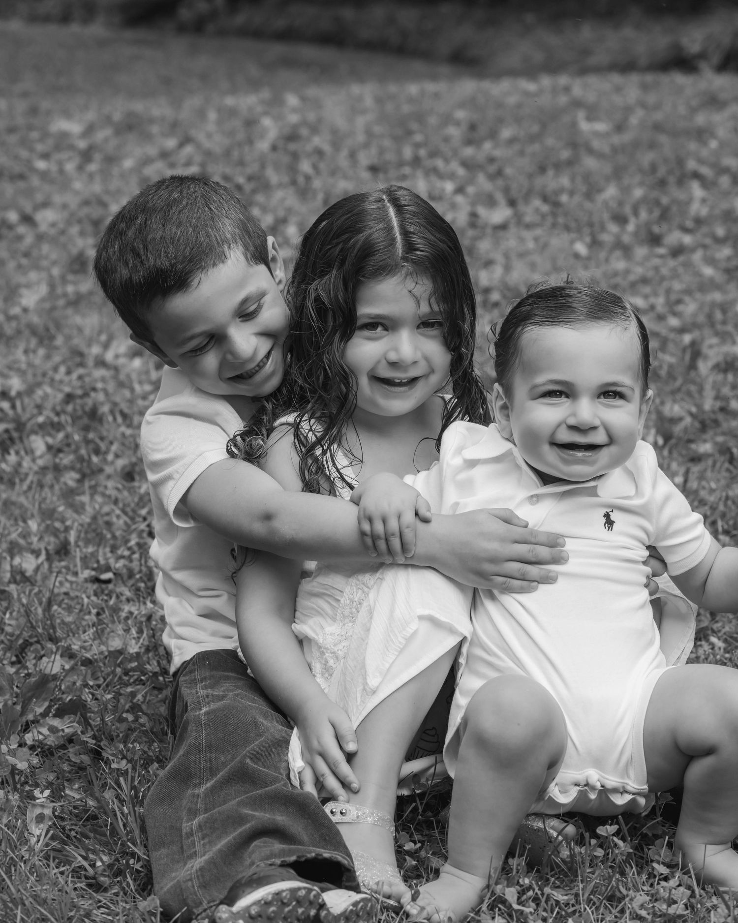 Black and white photo of three young siblings sitting together on grass, smiling and hugging outdoors.