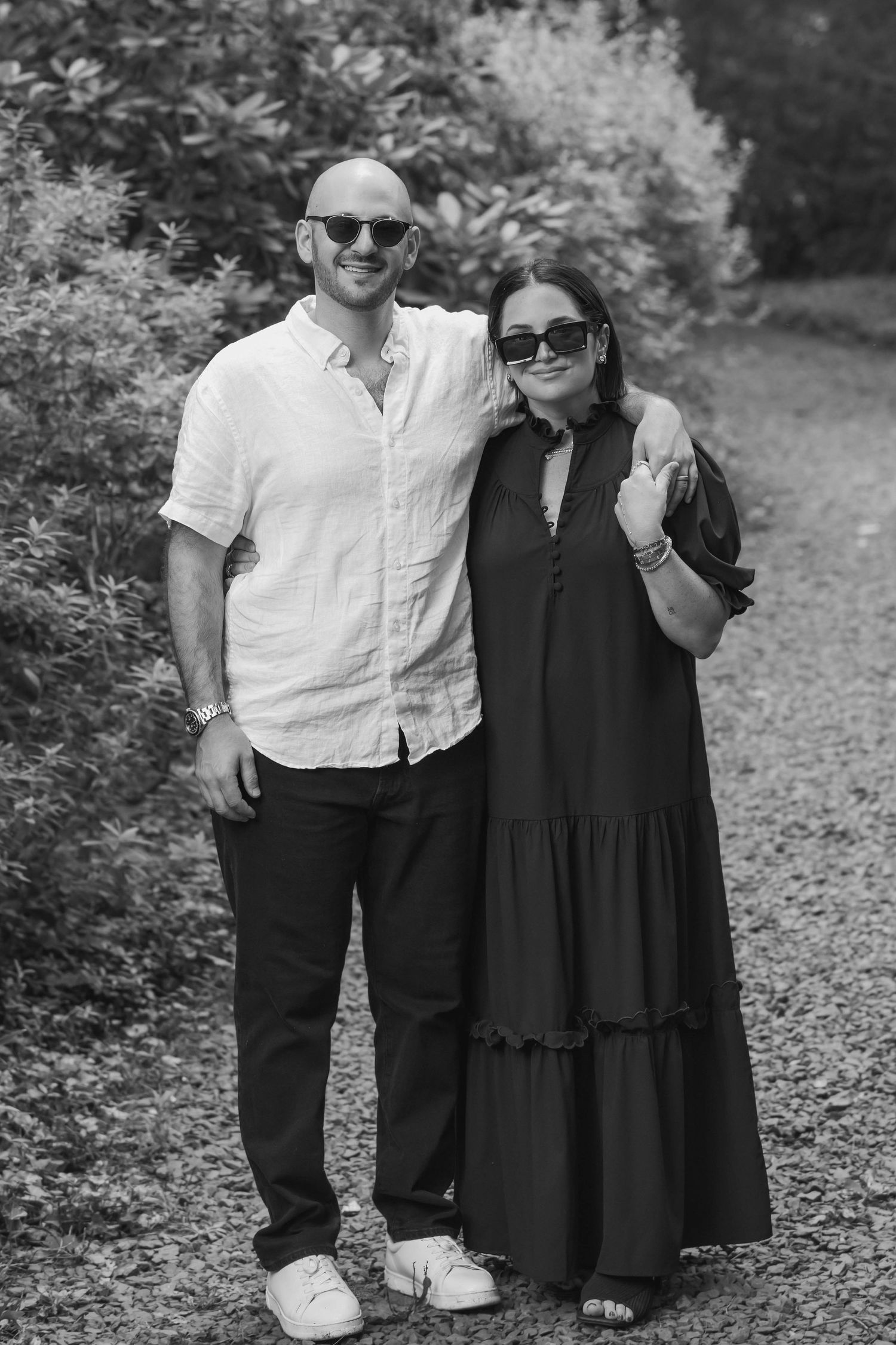 Black and white photo of a smiling couple in sunglasses posing outdoors on a garden path.