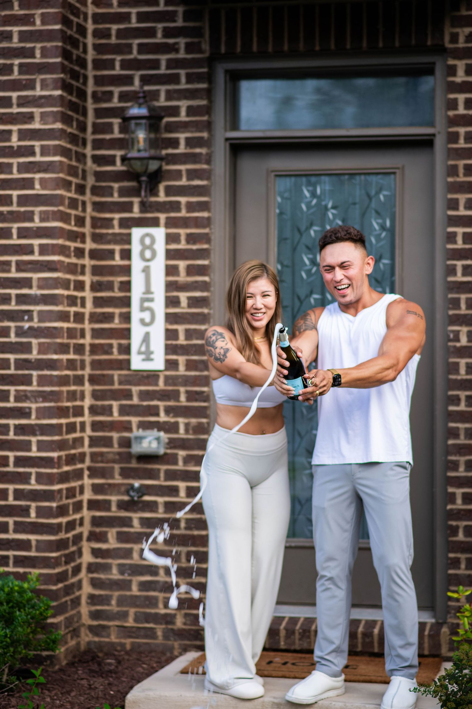 Happy couple celebrating new home purchase by popping champagne bottle in front of brick house door numbered 8154.