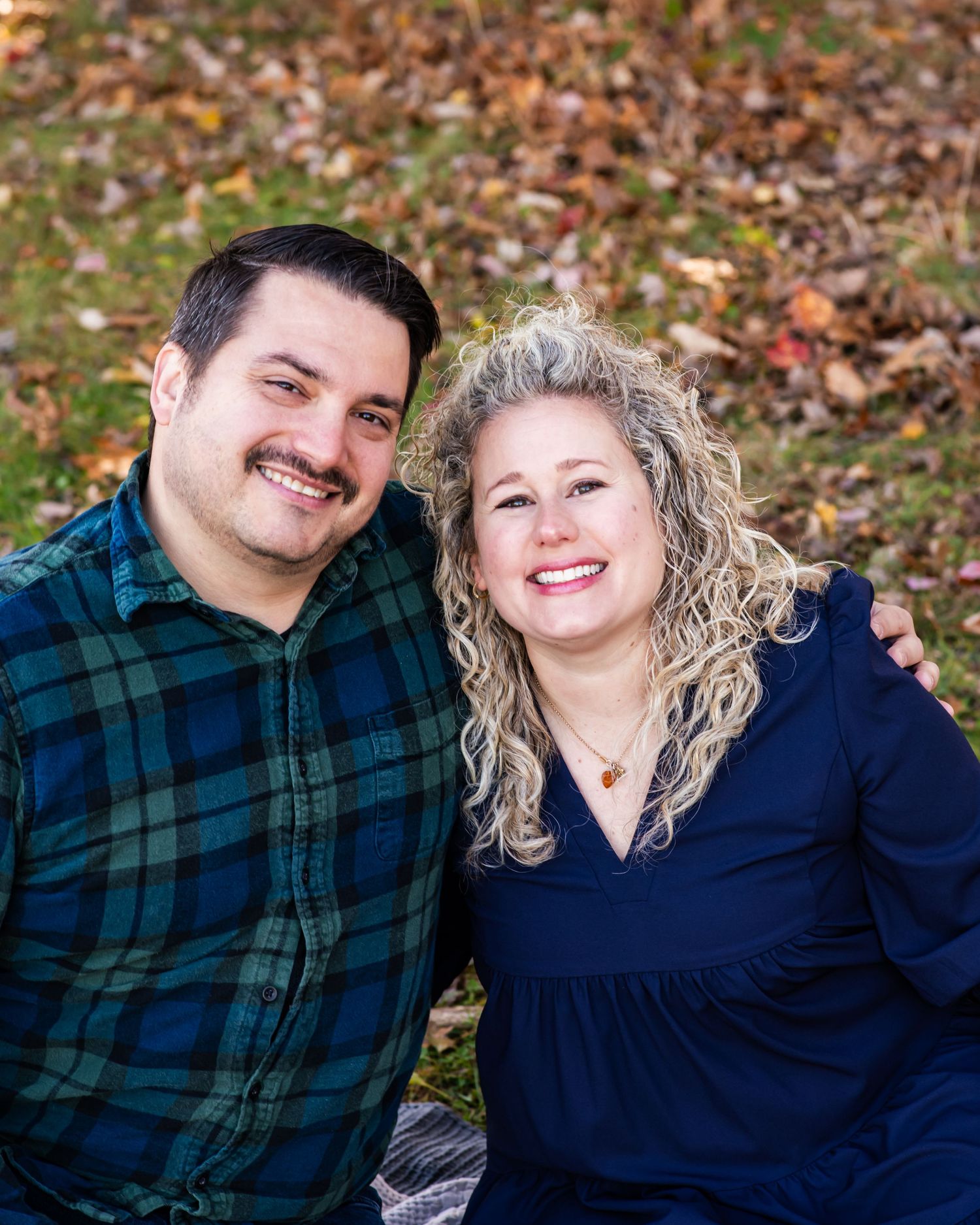 A couple sits together outdoors on fallen autumn leaves wearing coordinating navy and plaid outfits.
