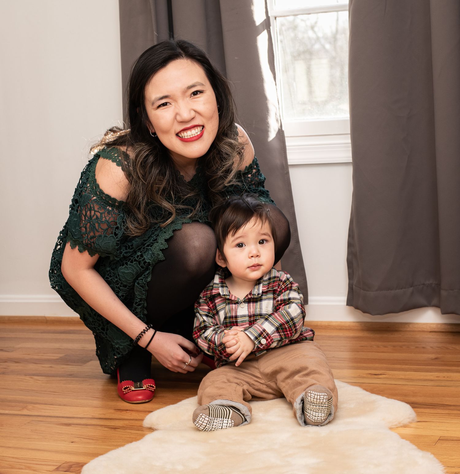 A woman in a green lace dress sits on a hardwood floor with a small child wearing plaid and khakis.