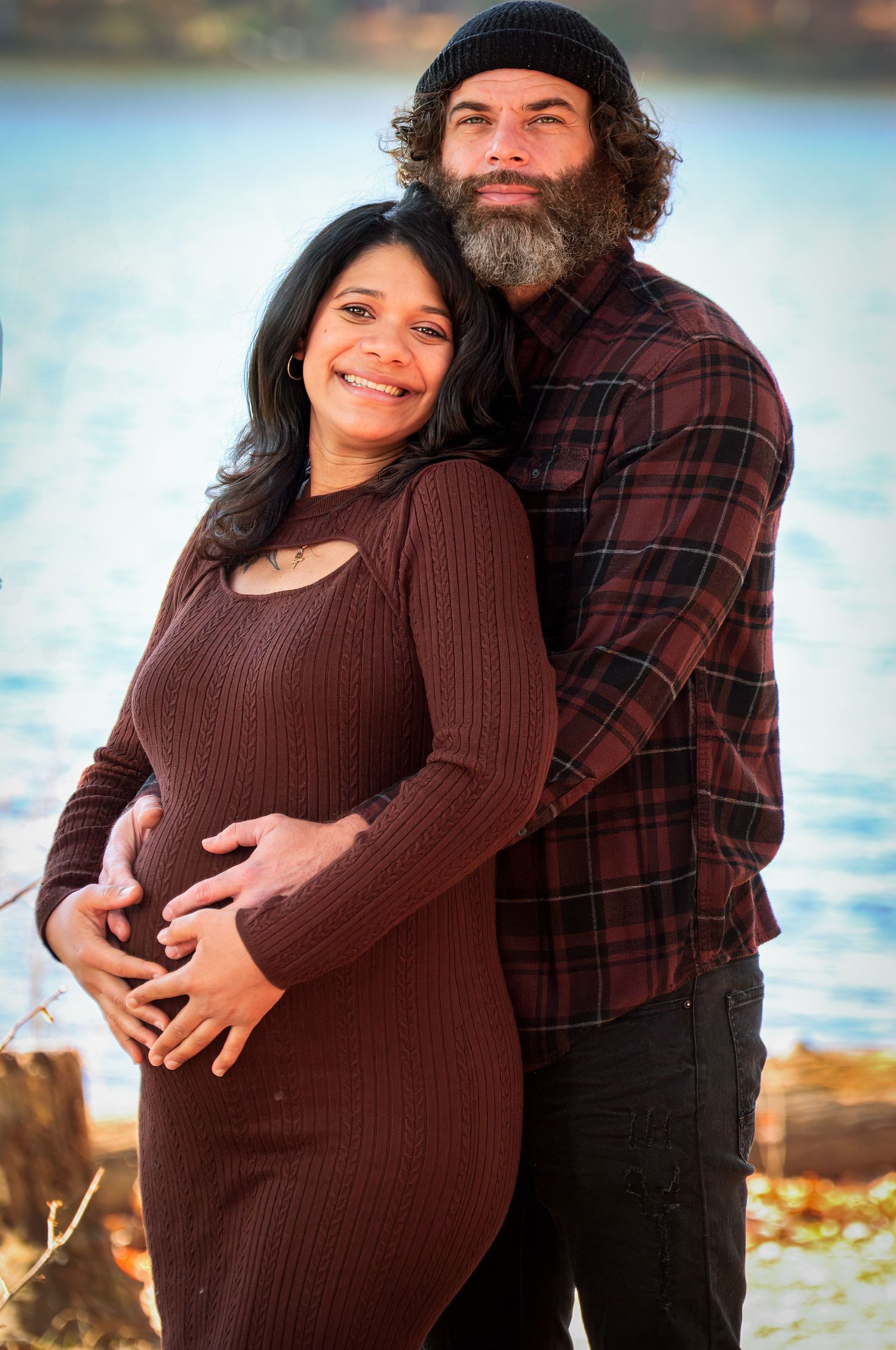 A couple poses together outdoors by the water, with the expectant mother wearing a brown sweater dress.
