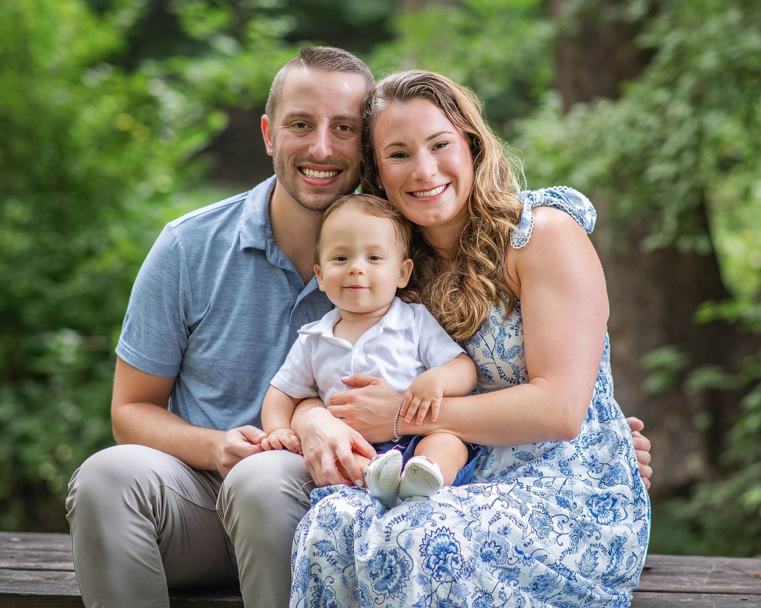Happy young family with smiling parents and baby boy posing outdoors on wooden bench surrounded by lush green trees.