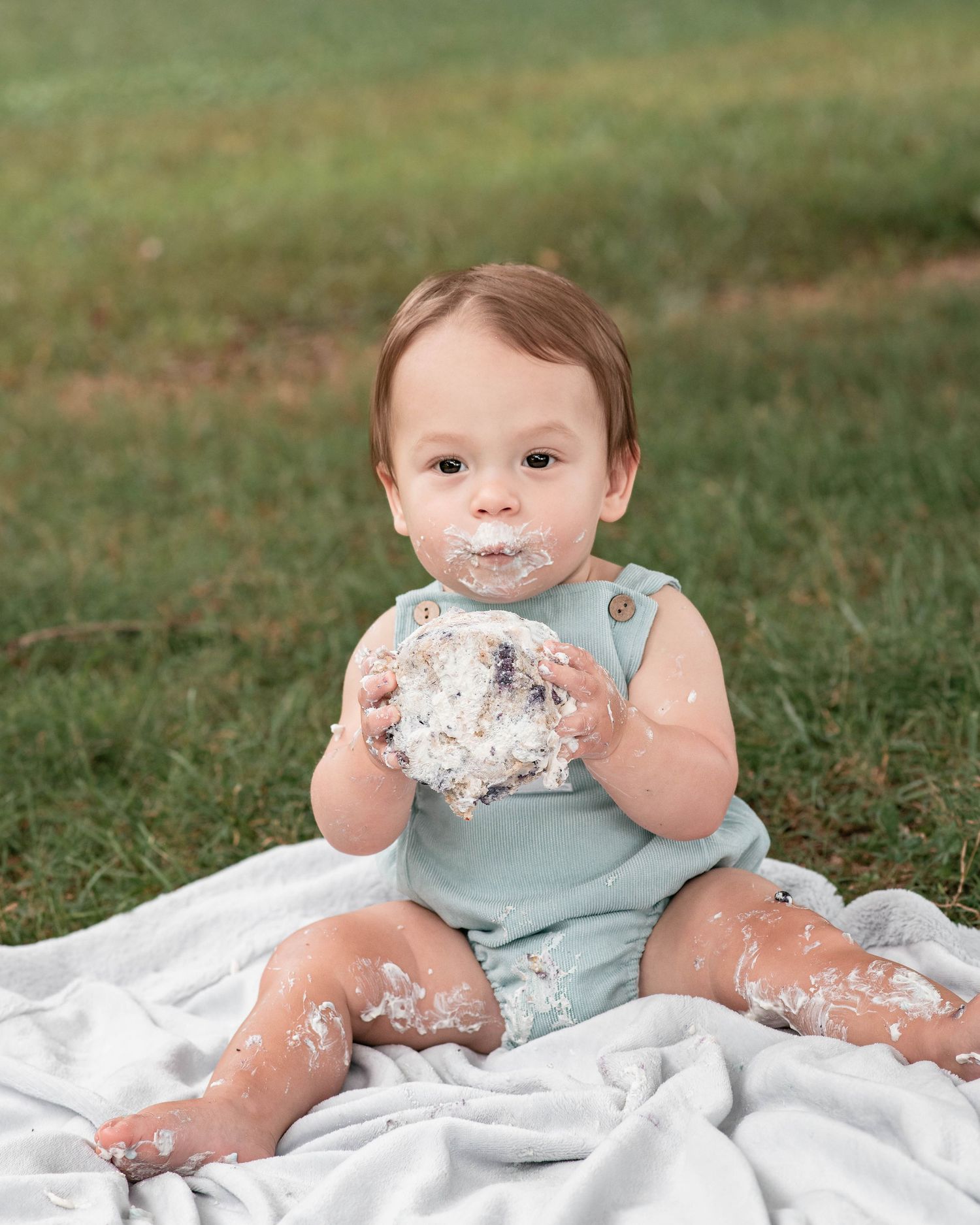 A baby in a mint romper smashes cake on a white blanket outdoors during a first birthday smash cake photoshoot.