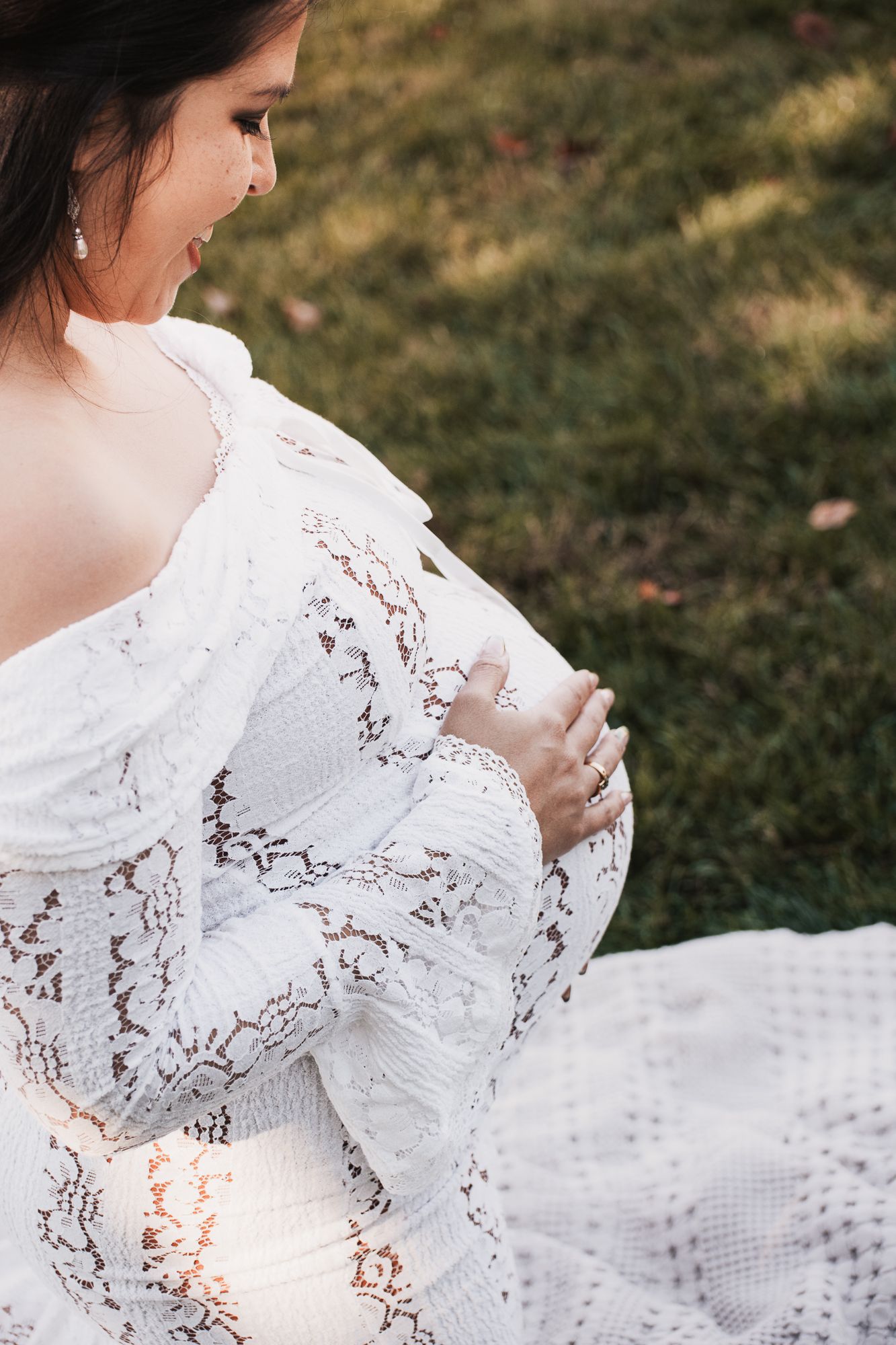 Pregnant woman in white lace dress sitting on grass, cradling her baby bump with both hands.