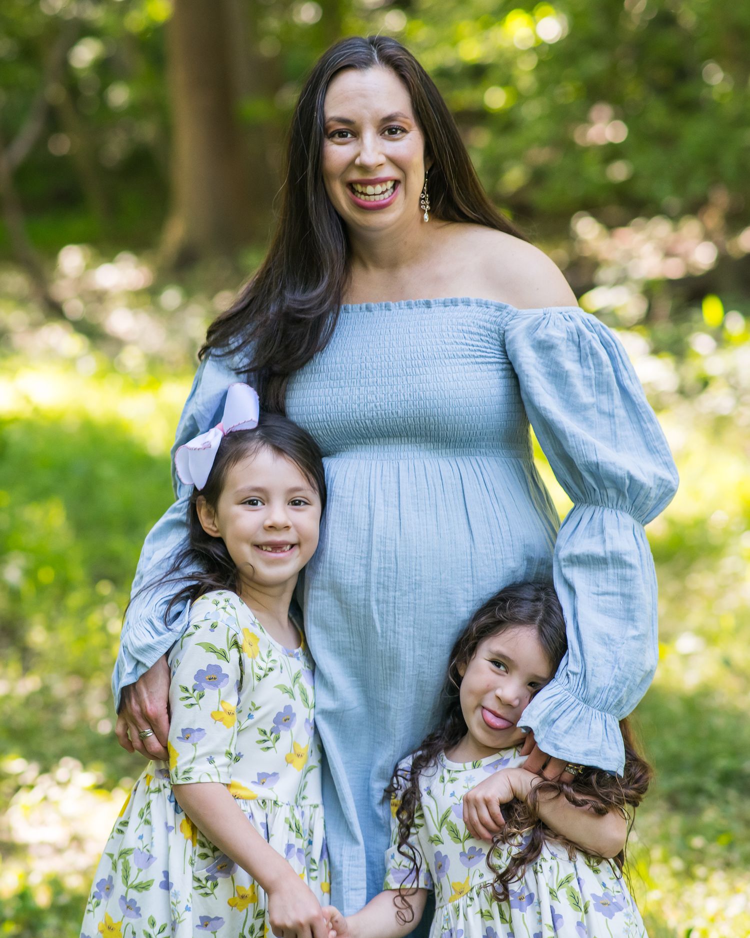 Pregnant woman in blue smocked dress poses outdoors with two young girls in floral dresses among green trees.