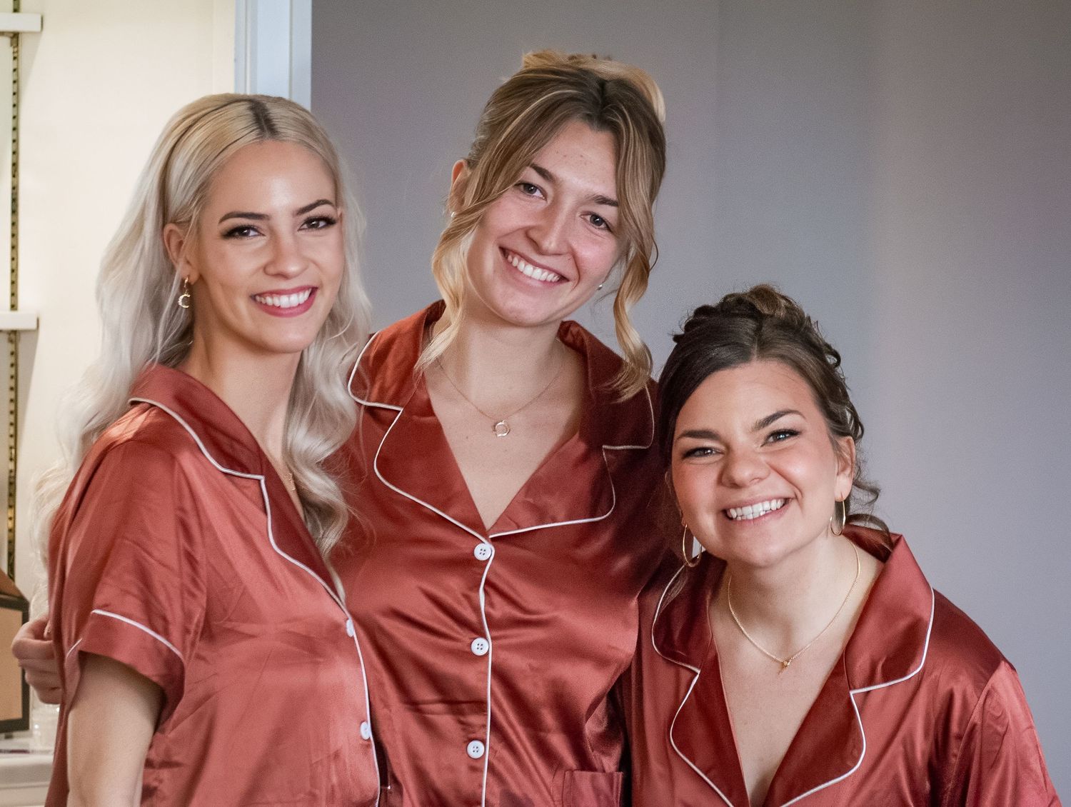 Three people wearing matching rust-colored satin pajama sets smile together in an indoor setting.