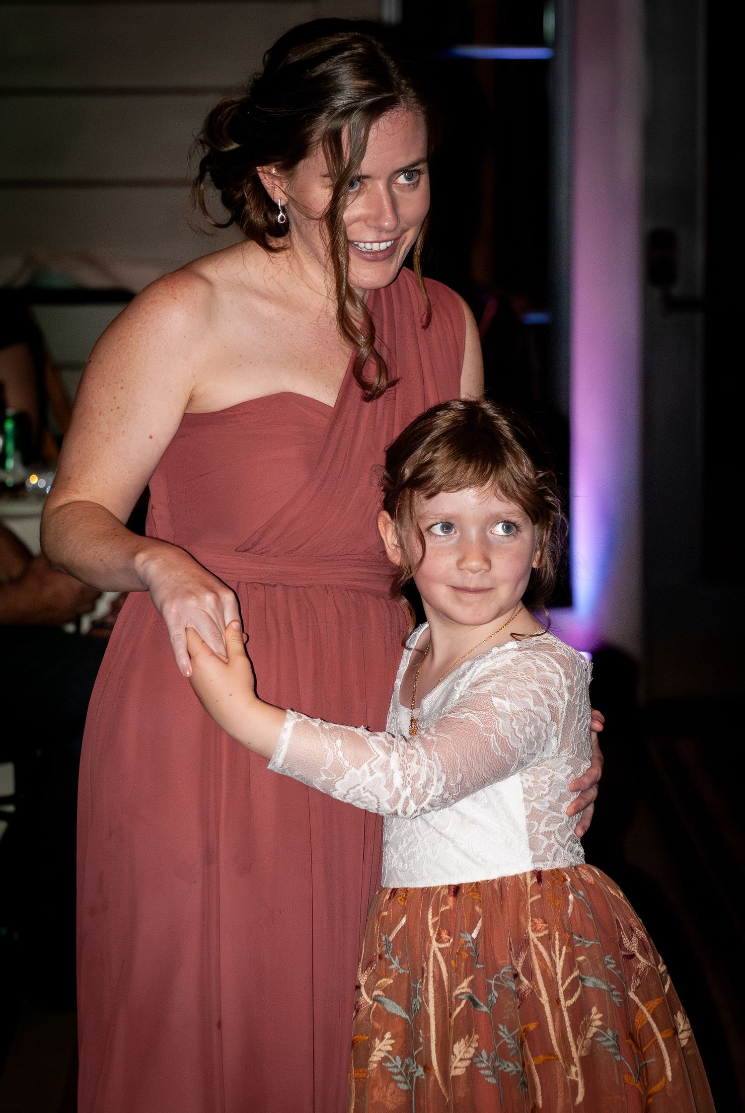 A young child in a white lace dress dances with someone wearing a dusty rose colored bridesmaid gown at a wedding reception.