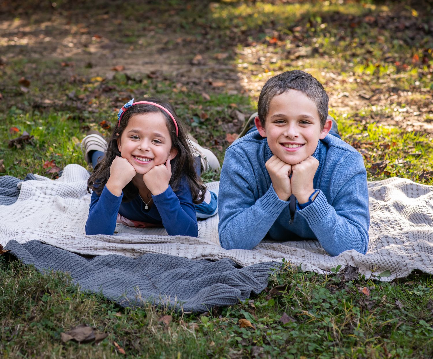 Two smiling children lying on a blanket in a park, posing with chins resting on their hands outdoors.