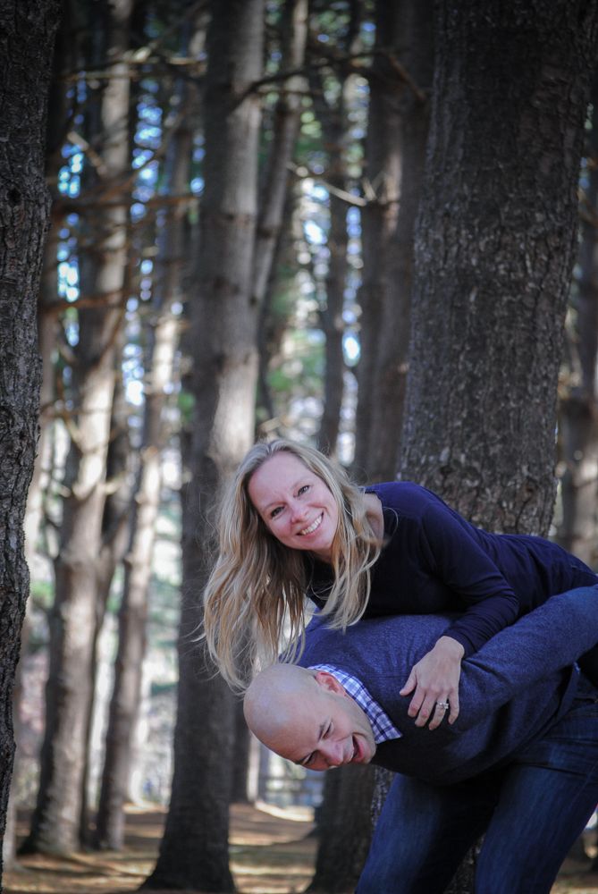 A woman smiles on a man's back during a playful piggyback ride among tall trees in a forest setting.