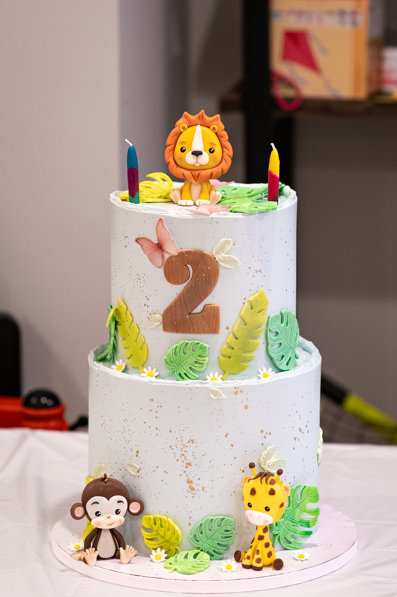 Two-tier safari birthday cake with fondant lion, monkey, giraffe, tropical leaves, and number 2 decoration with colorful candles.