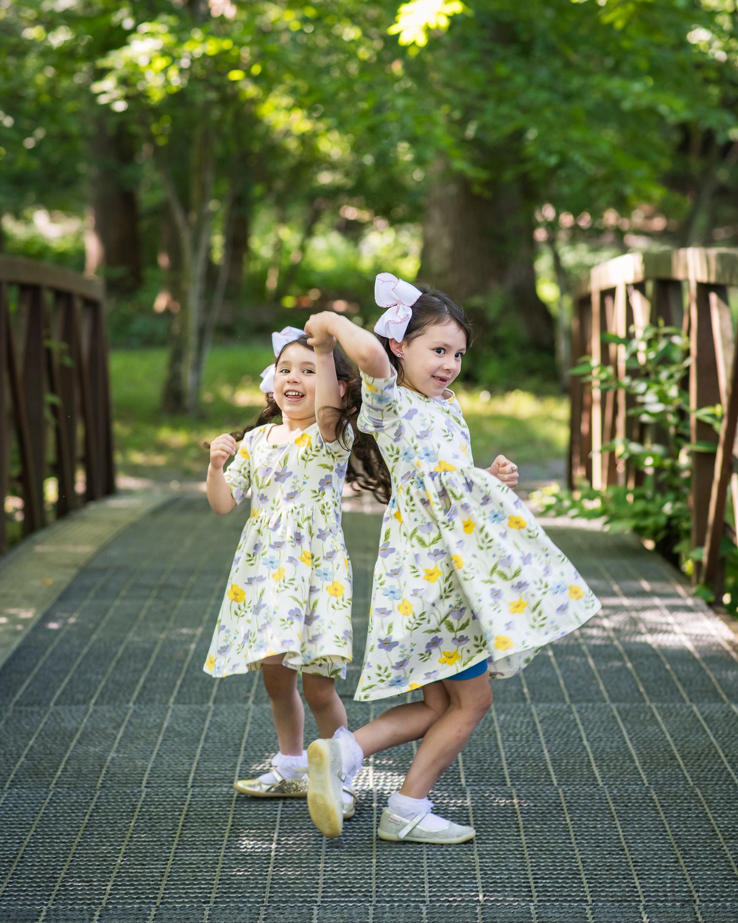 Two young girls in matching floral yellow dresses and white bows twirl on a garden bridge surrounded by lush green trees.