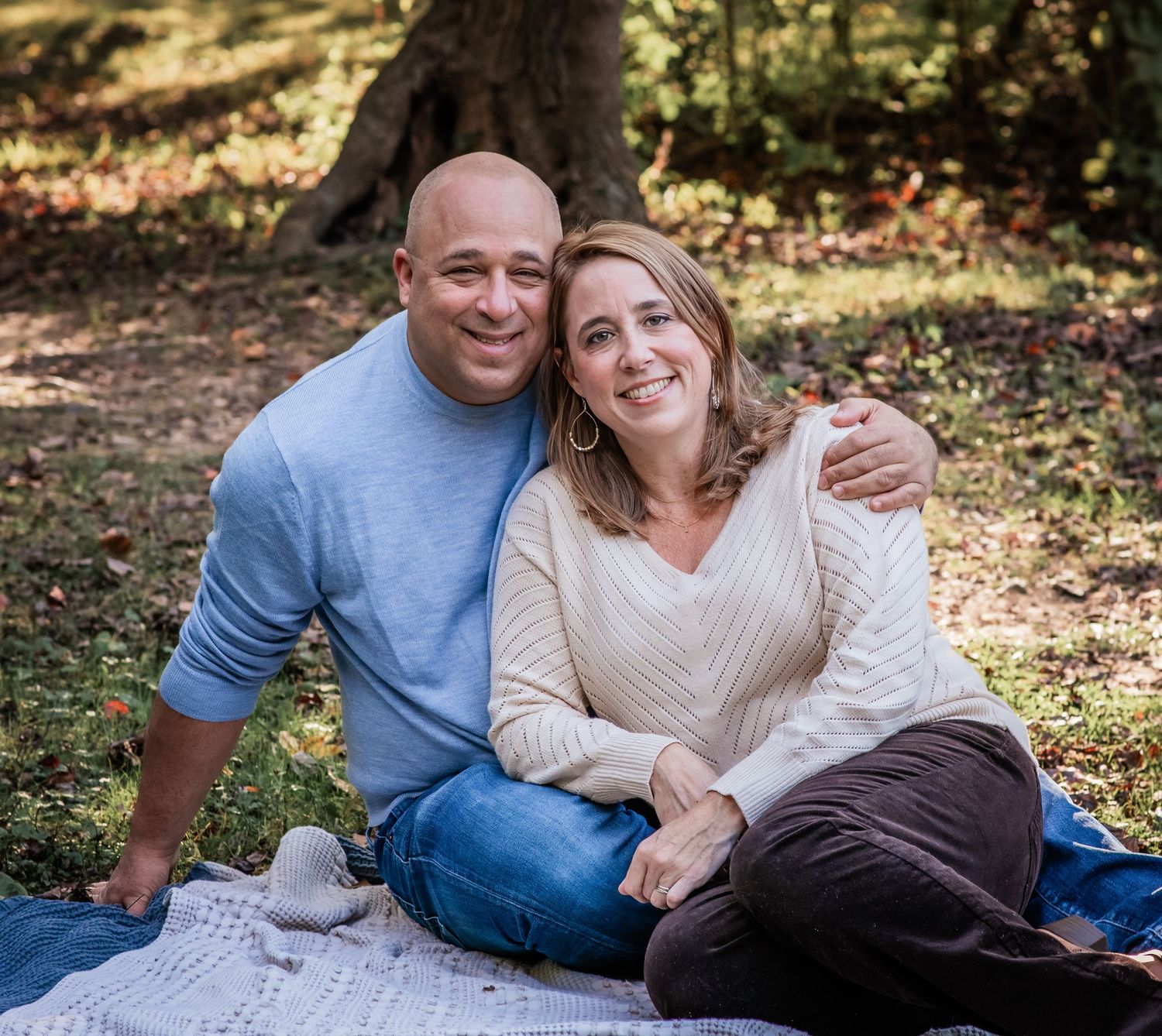 A couple sits together on a blanket in a wooded outdoor setting during a casual photo session.