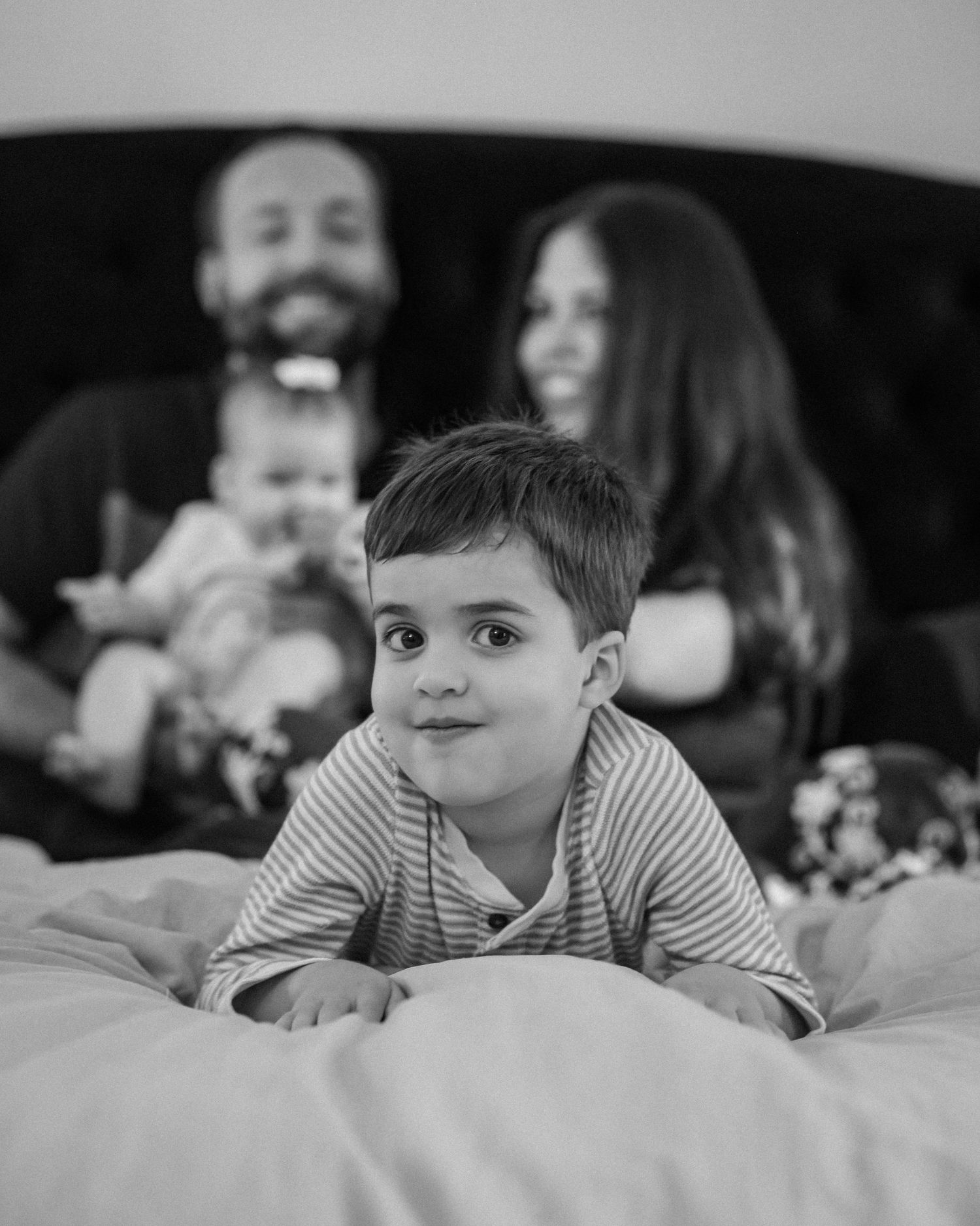 Black and white family photo with toddler boy lying on bed, parents smiling with baby in background.