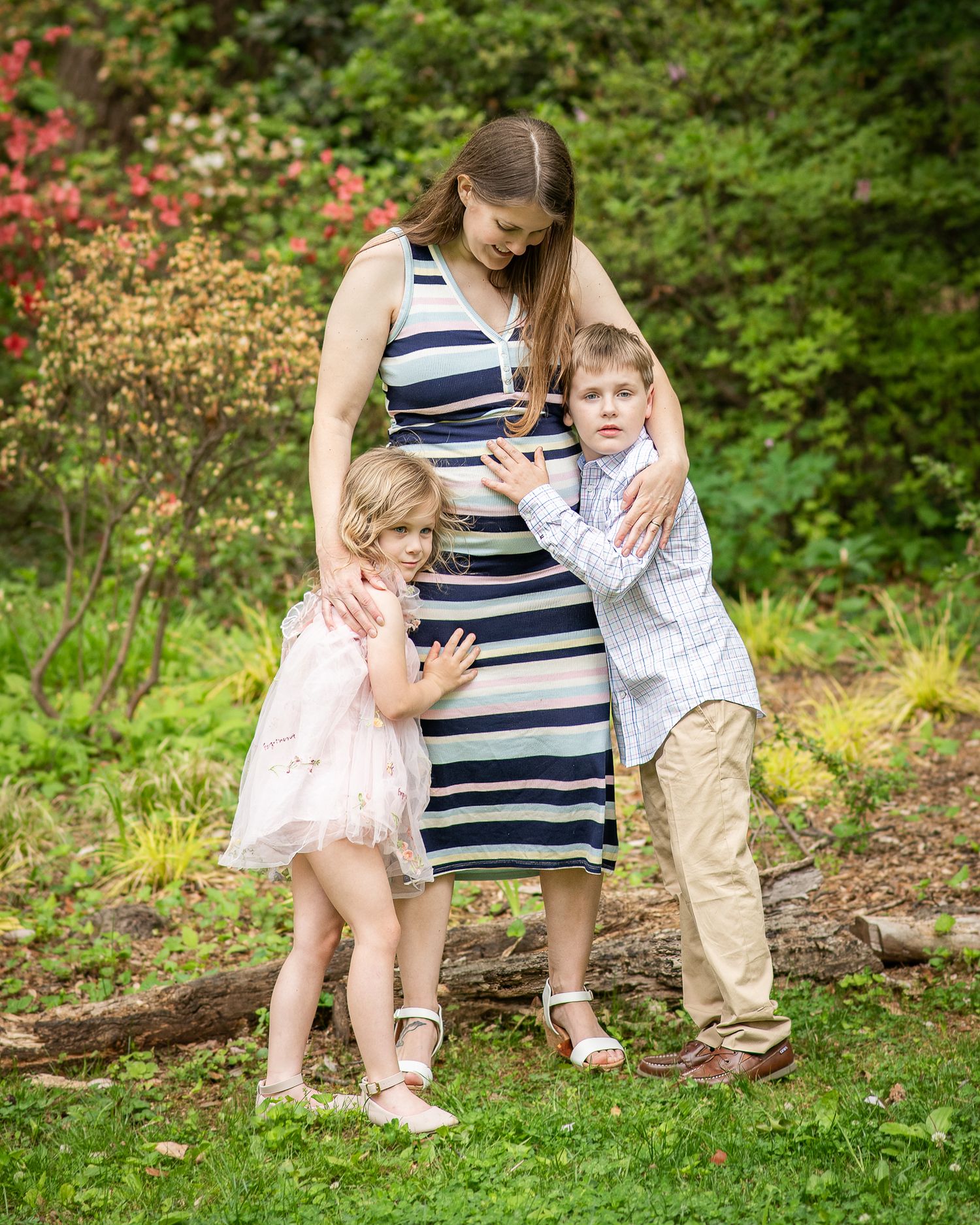 Pregnant woman in striped dress hugging two children in a lush garden setting during a maternity photo shoot.