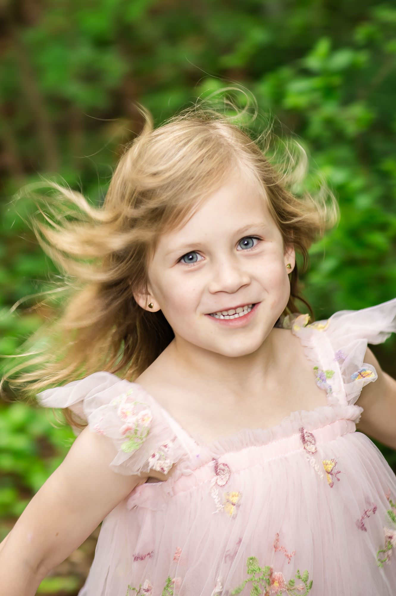 Smiling blonde girl with blue eyes in pink floral dress, hair blowing in wind, outdoors with green foliage background.