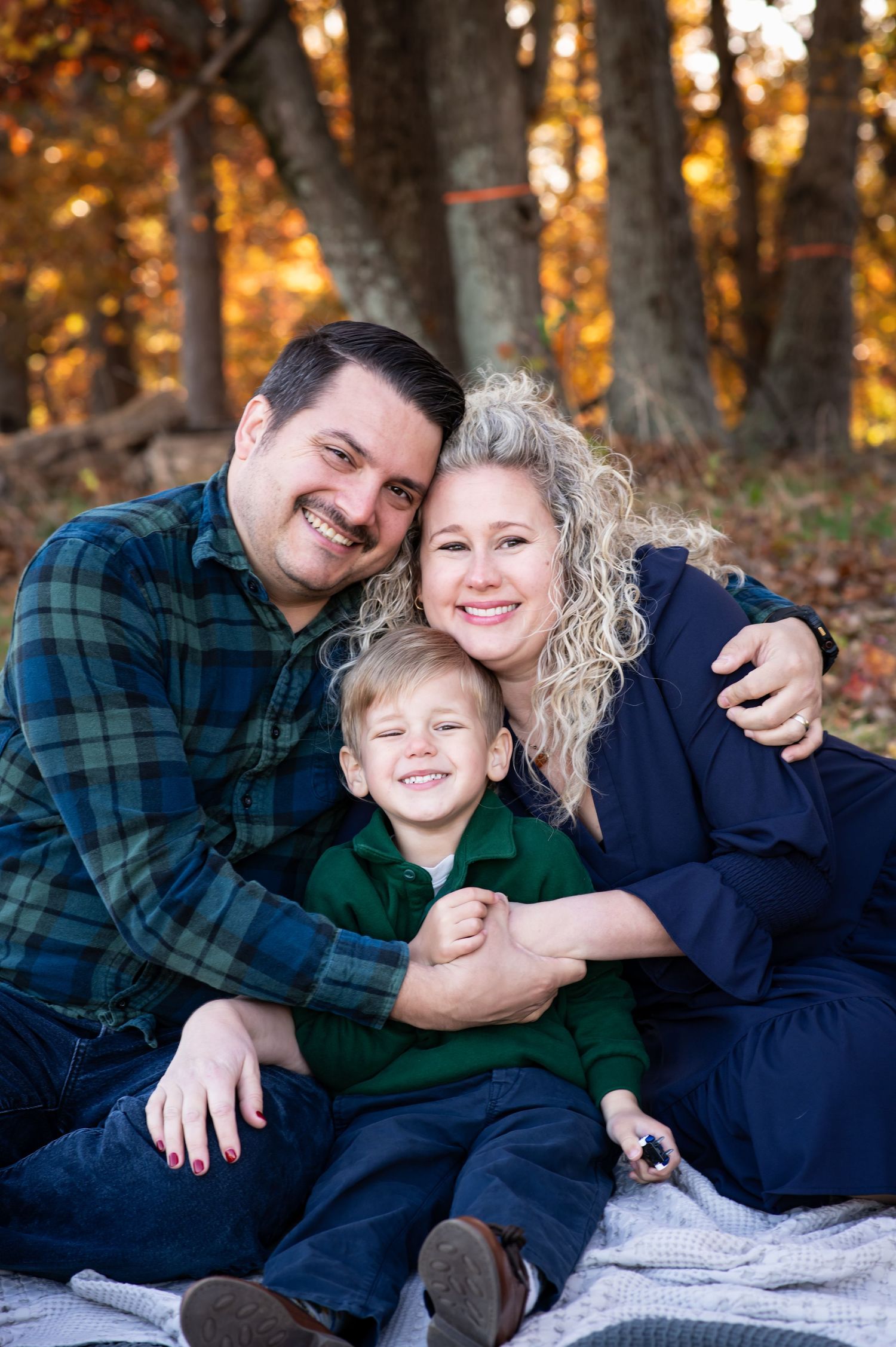 Family enjoying playful moments together on a blanket in autumn woods with golden sunlight filtering through trees.
