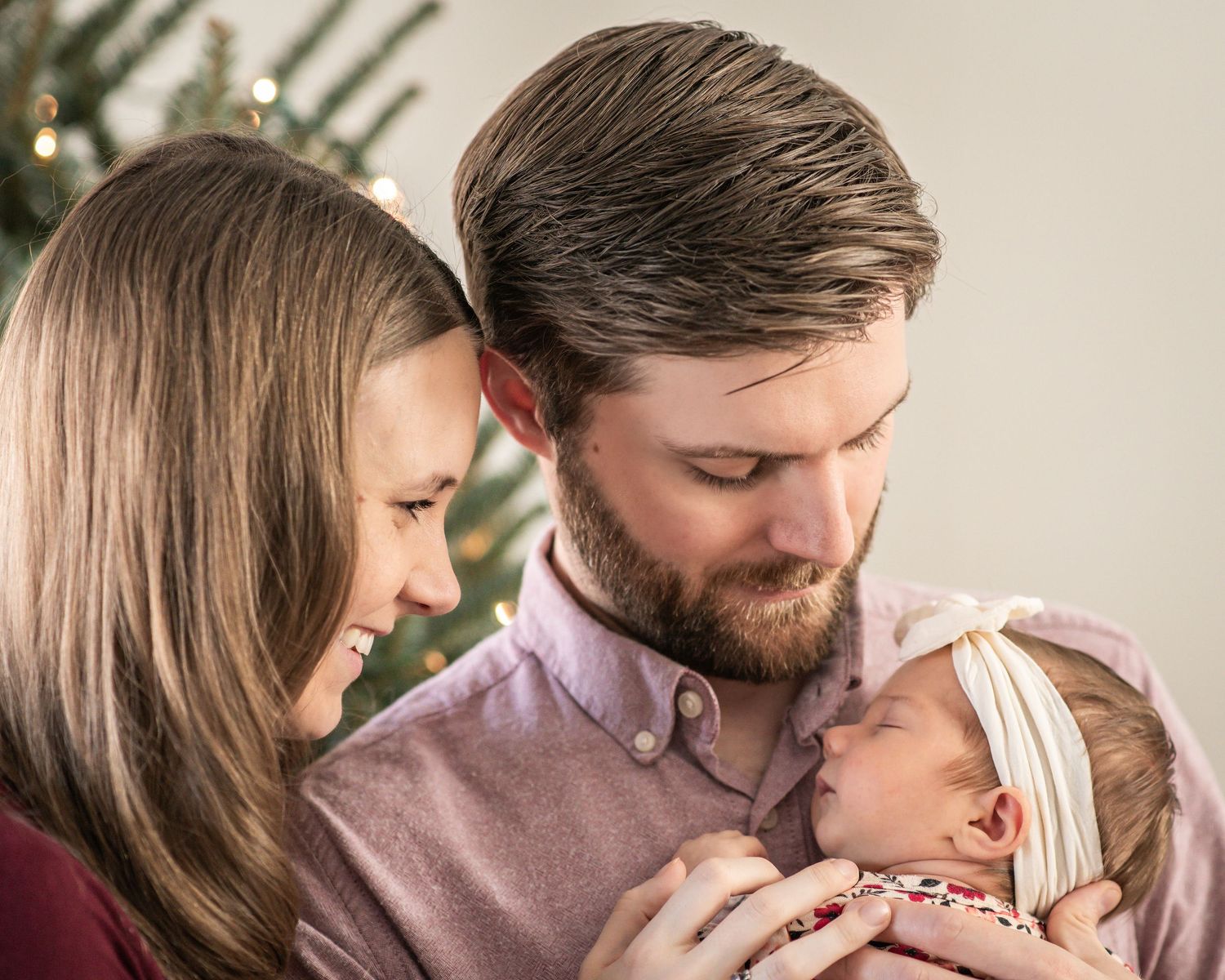 Young couple gazing lovingly at their newborn baby near a Christmas tree in a warm family portrait.