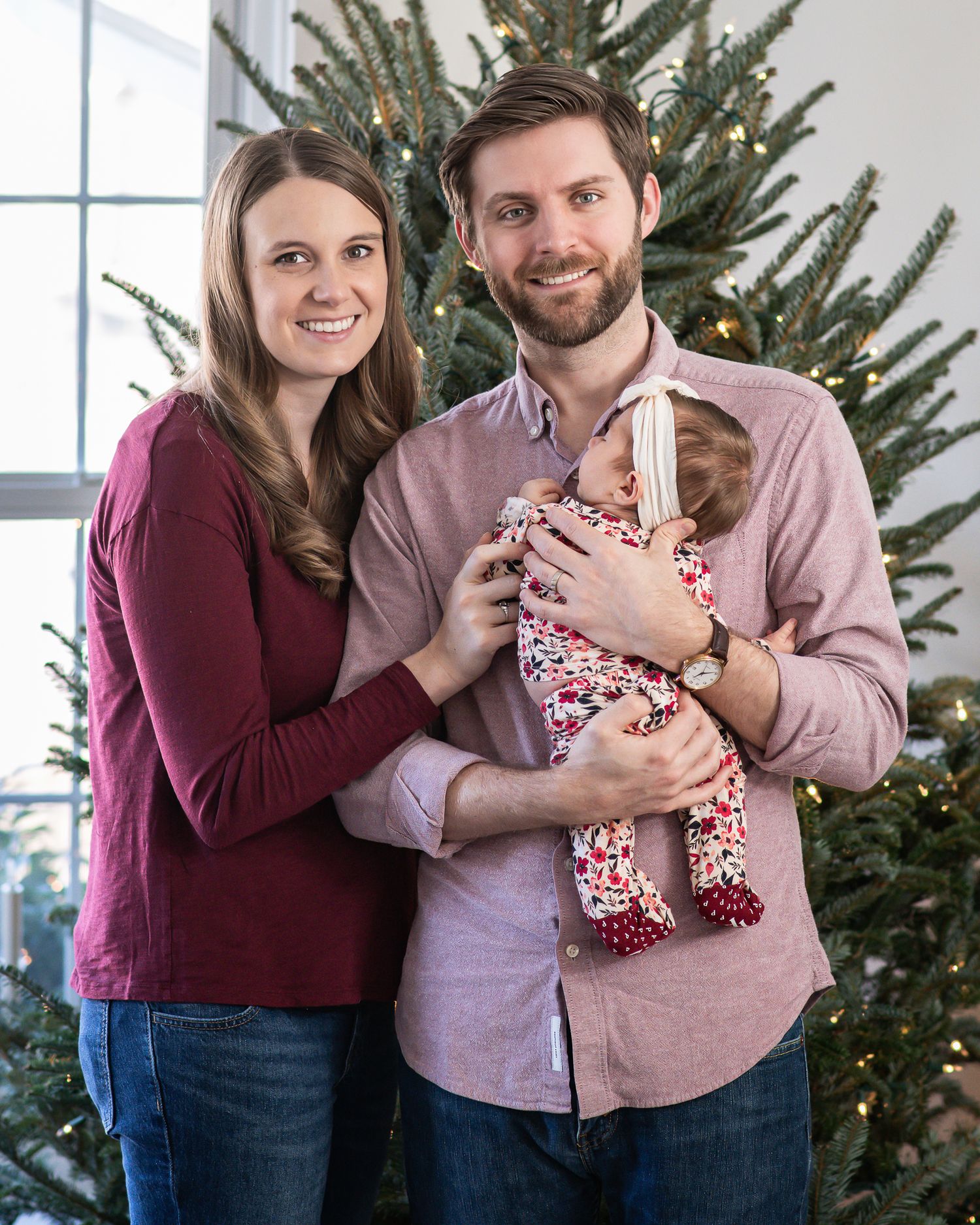 A couple in burgundy and pink sweaters poses together with a Christmas tree in the background.