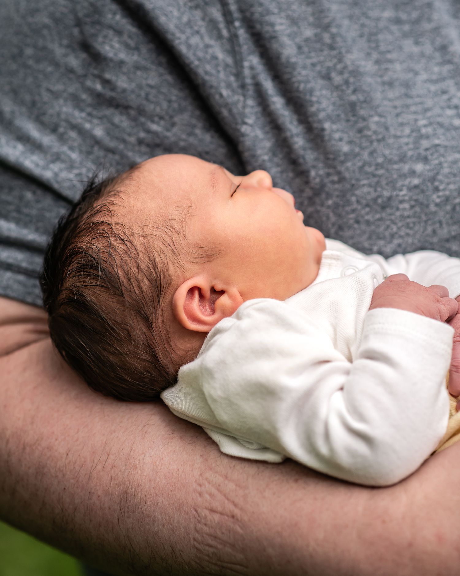Newborn baby sleeping peacefully on father's arm, dressed in white onesie, with dark hair and serene expression.