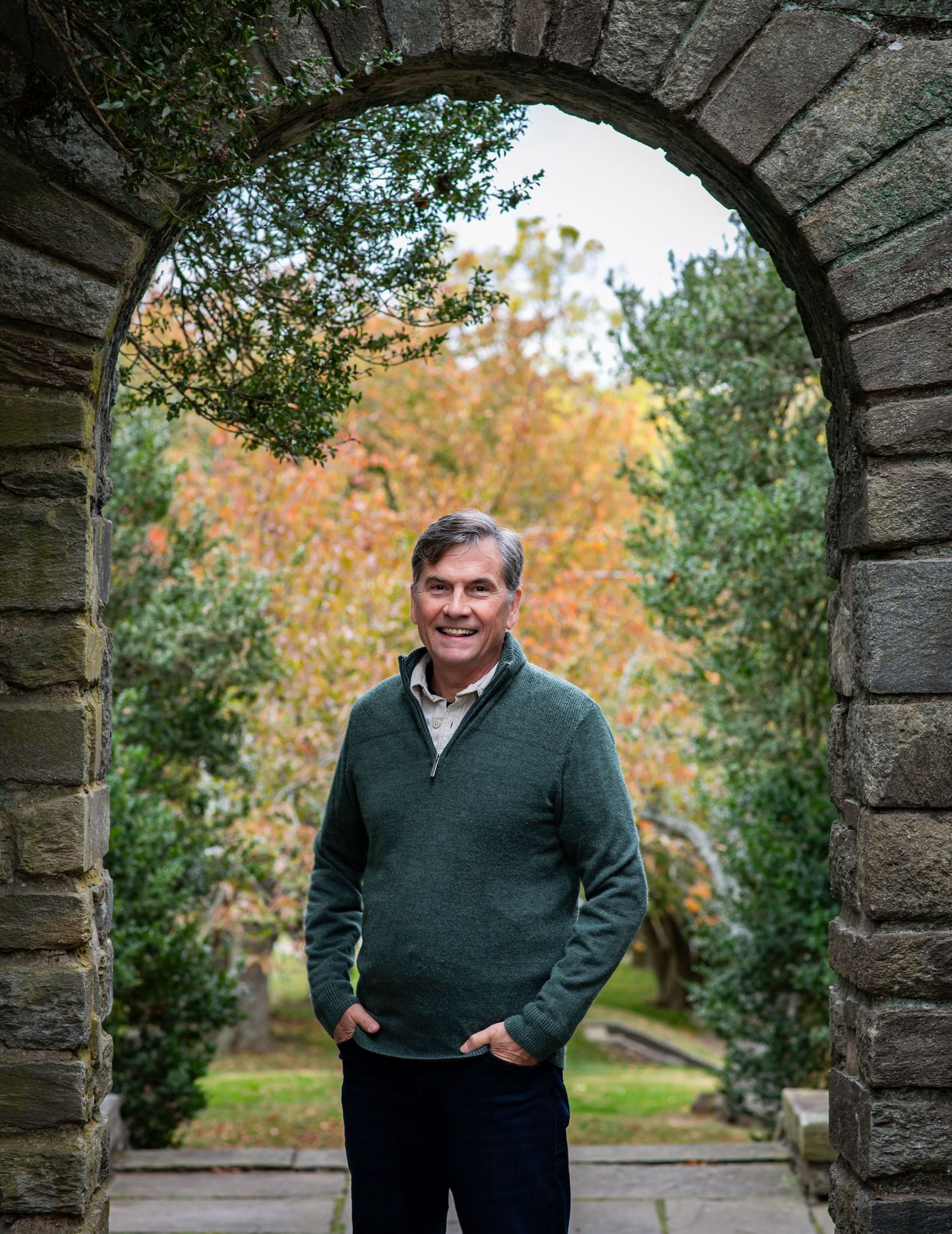 A person in a green sweater stands in a stone archway during autumn with trees and foliage in the background.