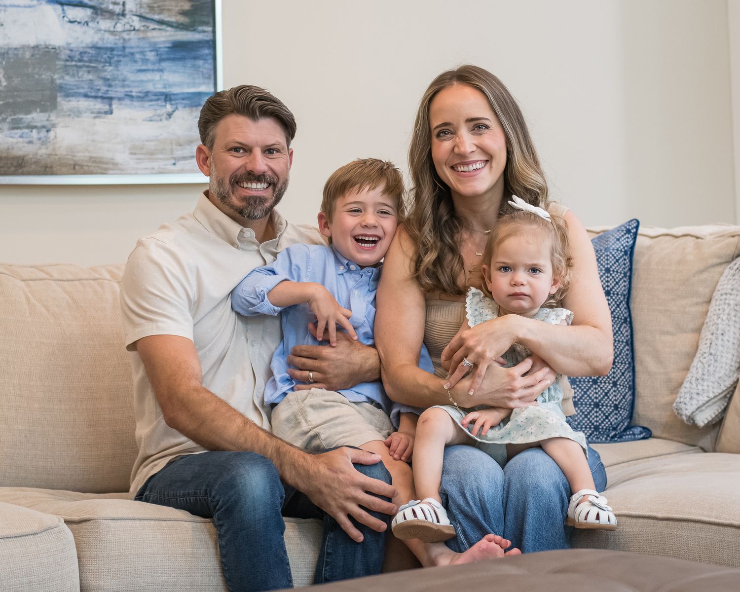 Happy family of four smiling together on a beige couch, parents with young boy and baby girl in a cozy home.