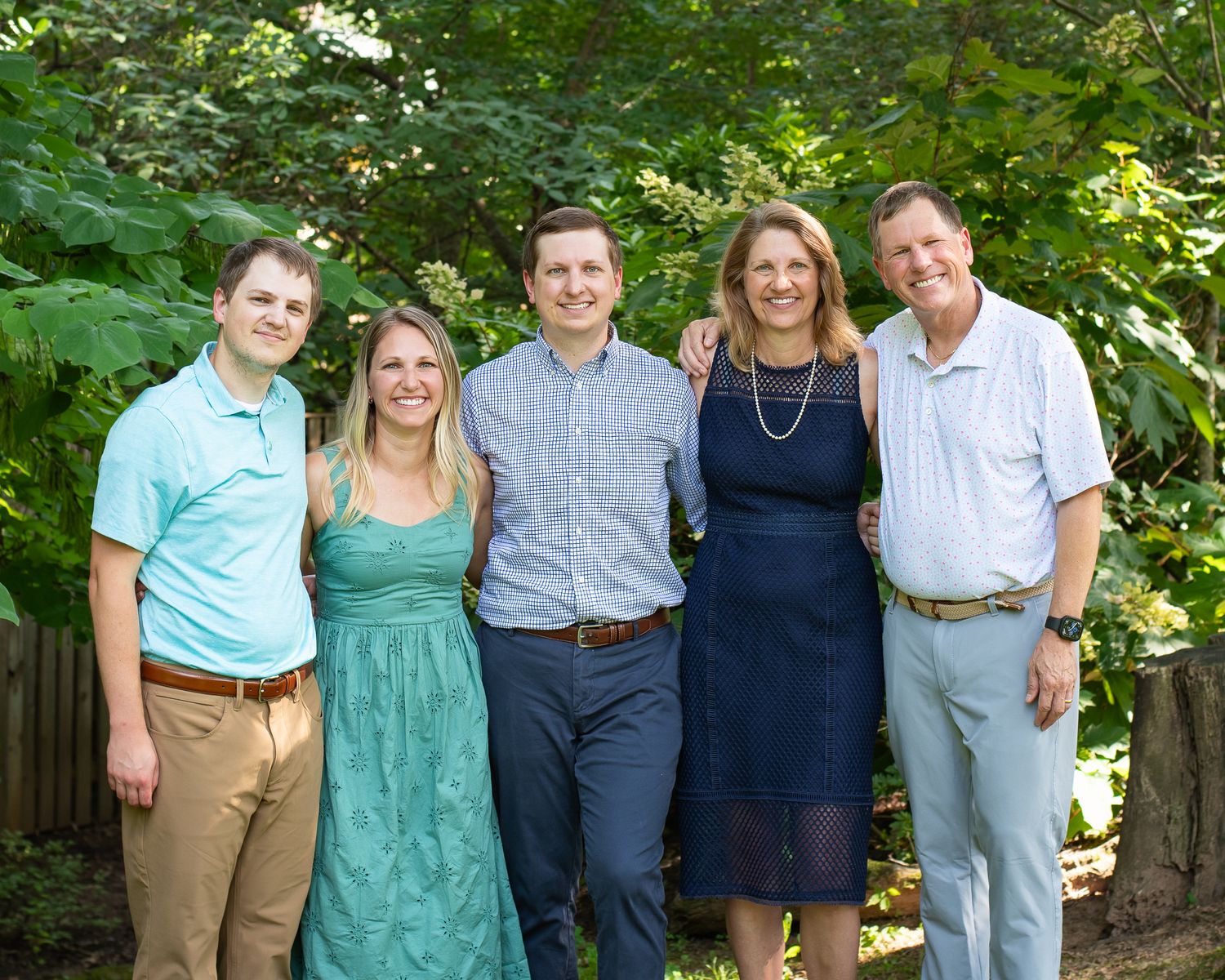 A smiling family of five poses outdoors among lush green trees in casual summer attire.