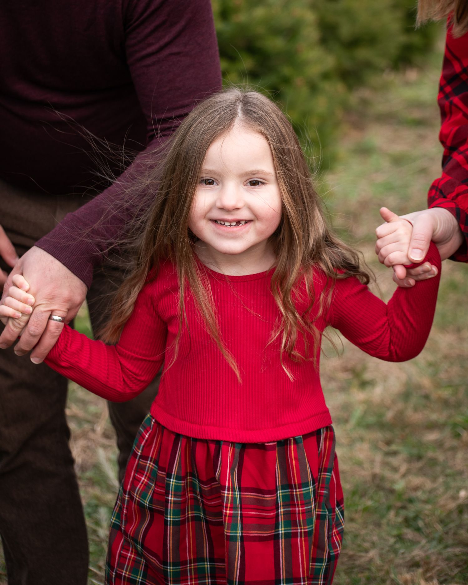 A smiling young girl in a red top and plaid skirt holds hands with adults outdoors in a festive holiday photo.