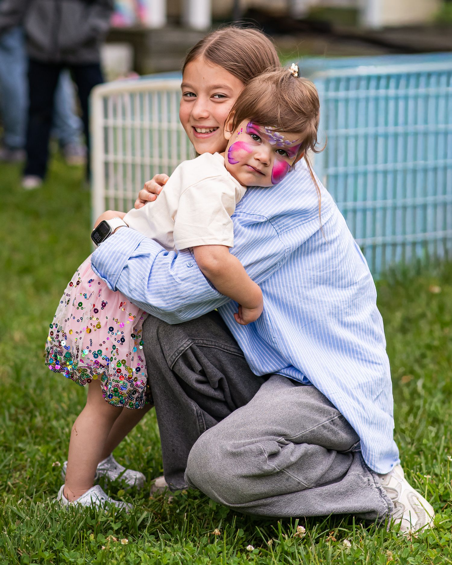 Two young girls hugging and smiling outdoors, one with colorful butterfly face paint, at a festive event.