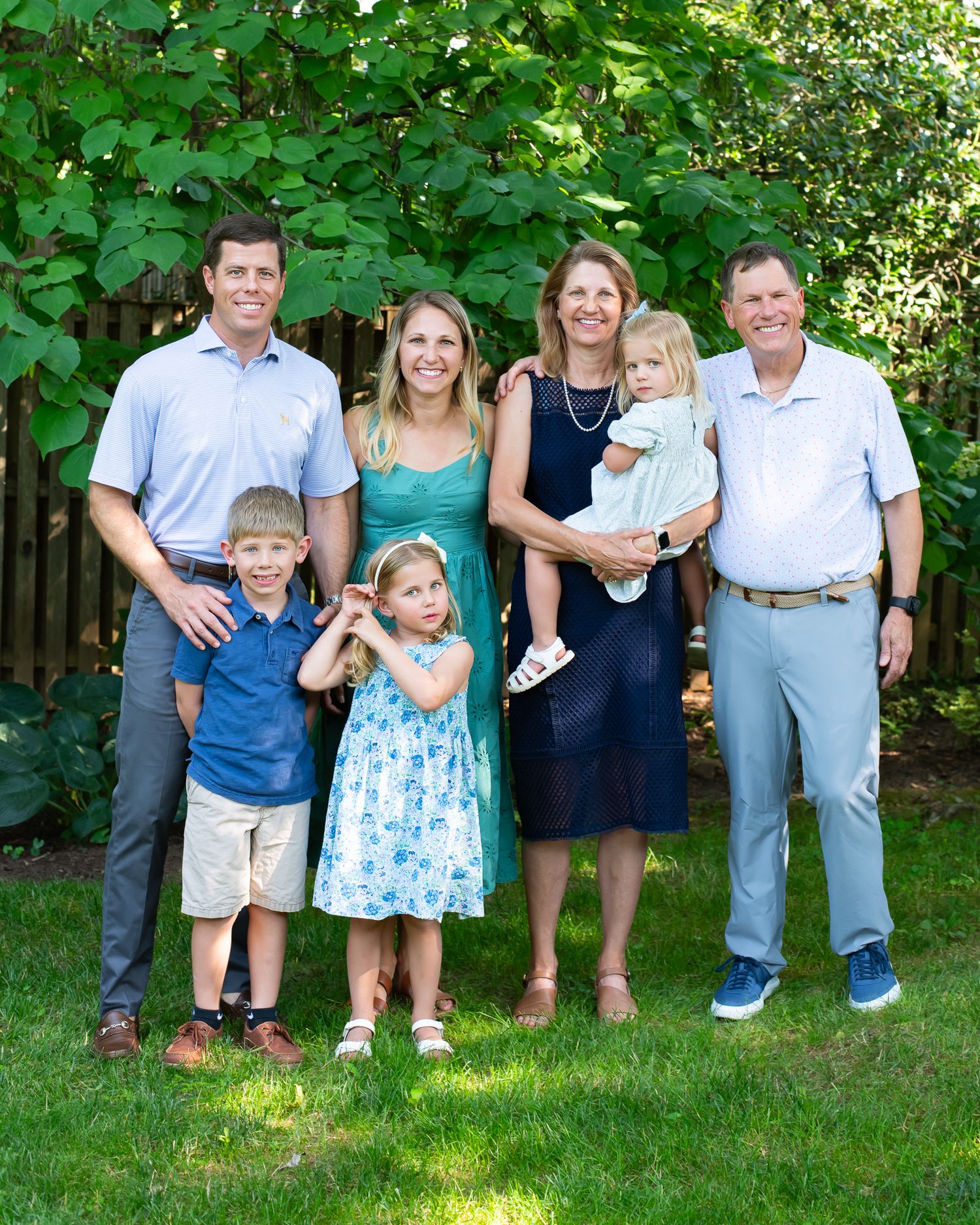 A happy family of seven poses together outdoors on a sunny day, smiling on a green lawn with lush foliage behind them.