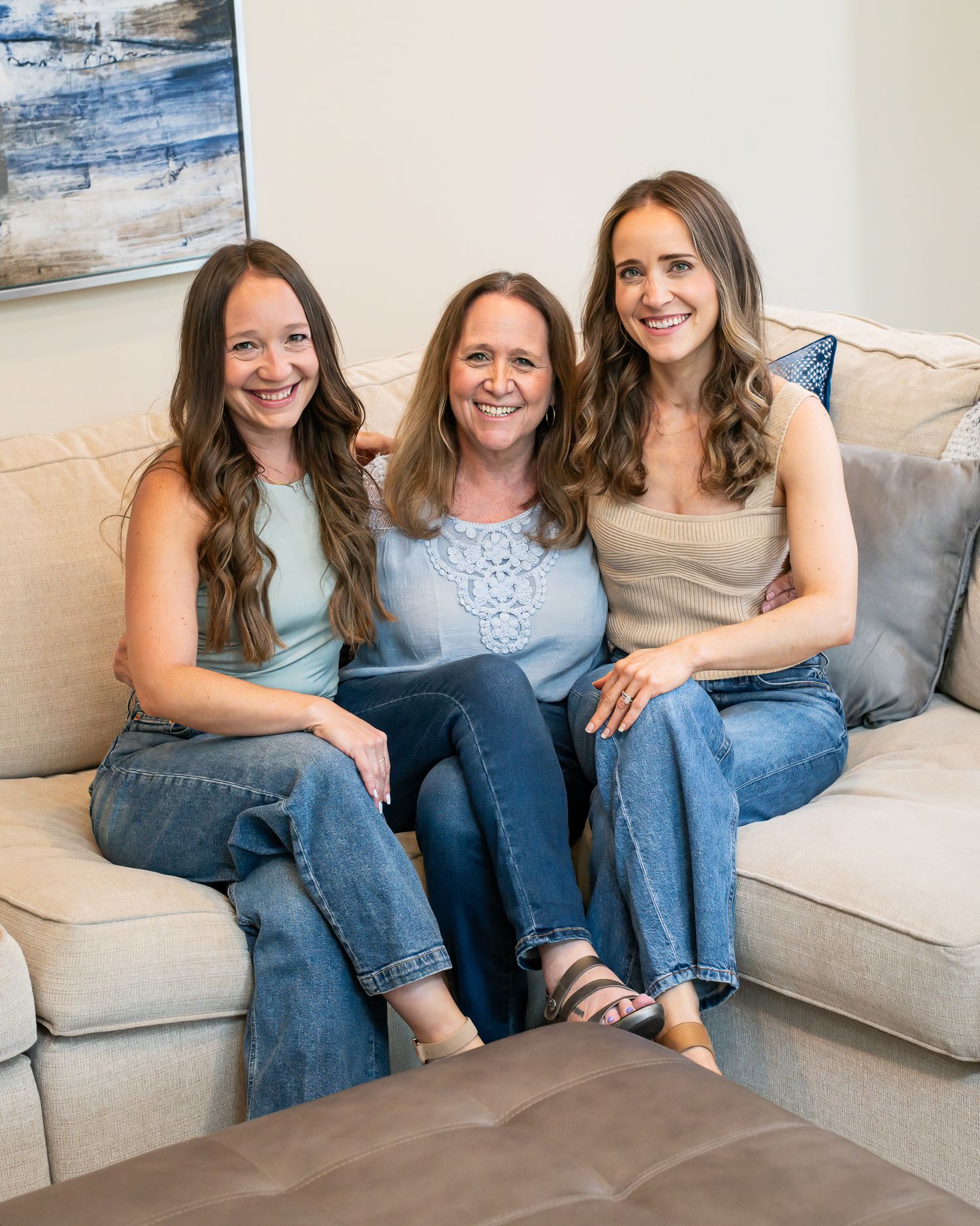 Three smiling women sitting together on a beige couch in a bright living room, wearing casual jeans and tops.