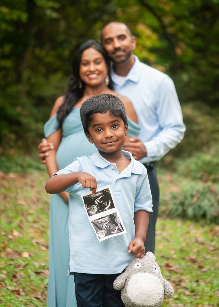 A family in coordinated light blue and white outfits poses together for outdoor pregnancy announcement photos.