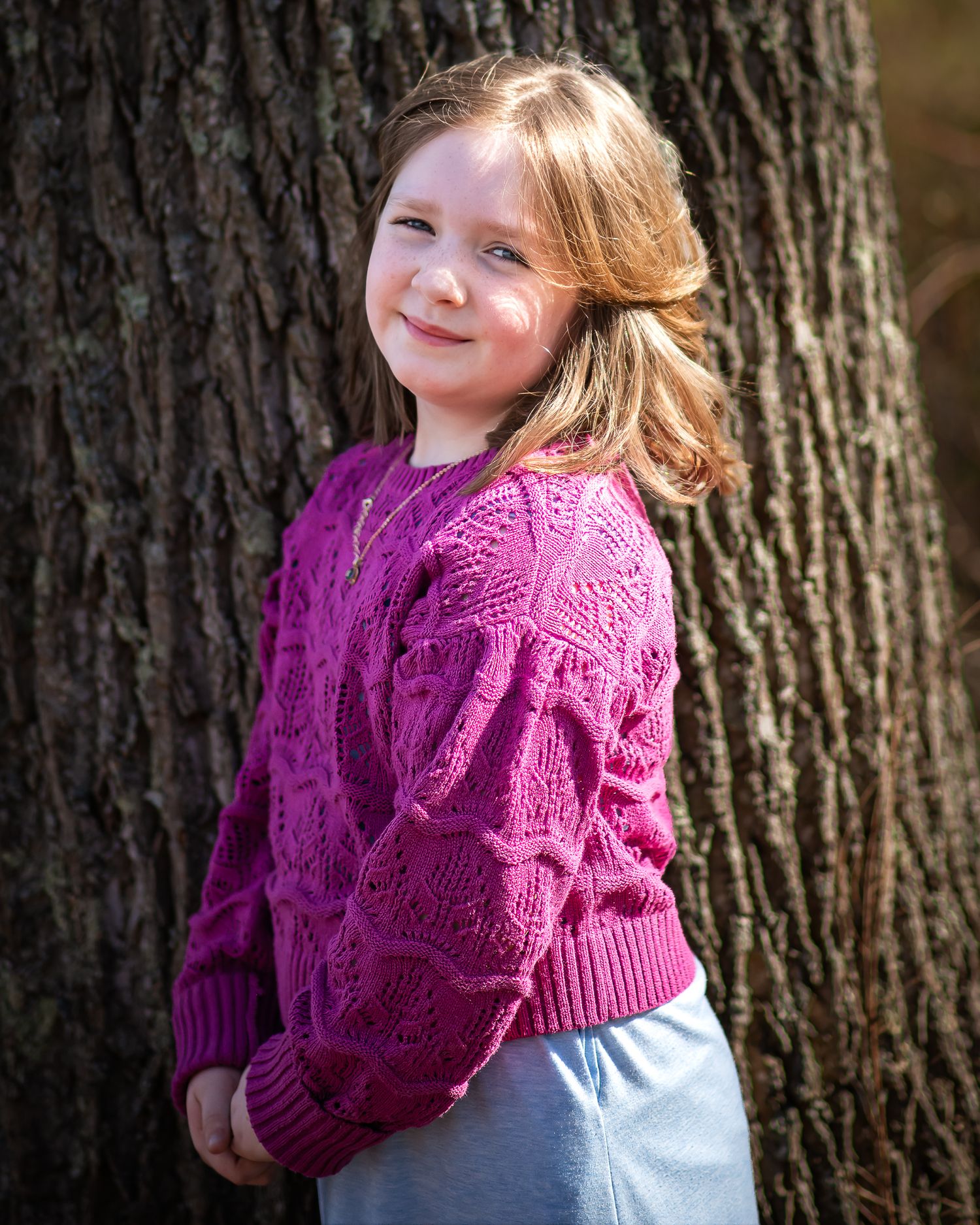 A young person in a bright magenta knit sweater stands smiling against a large tree trunk in natural outdoor lighting.