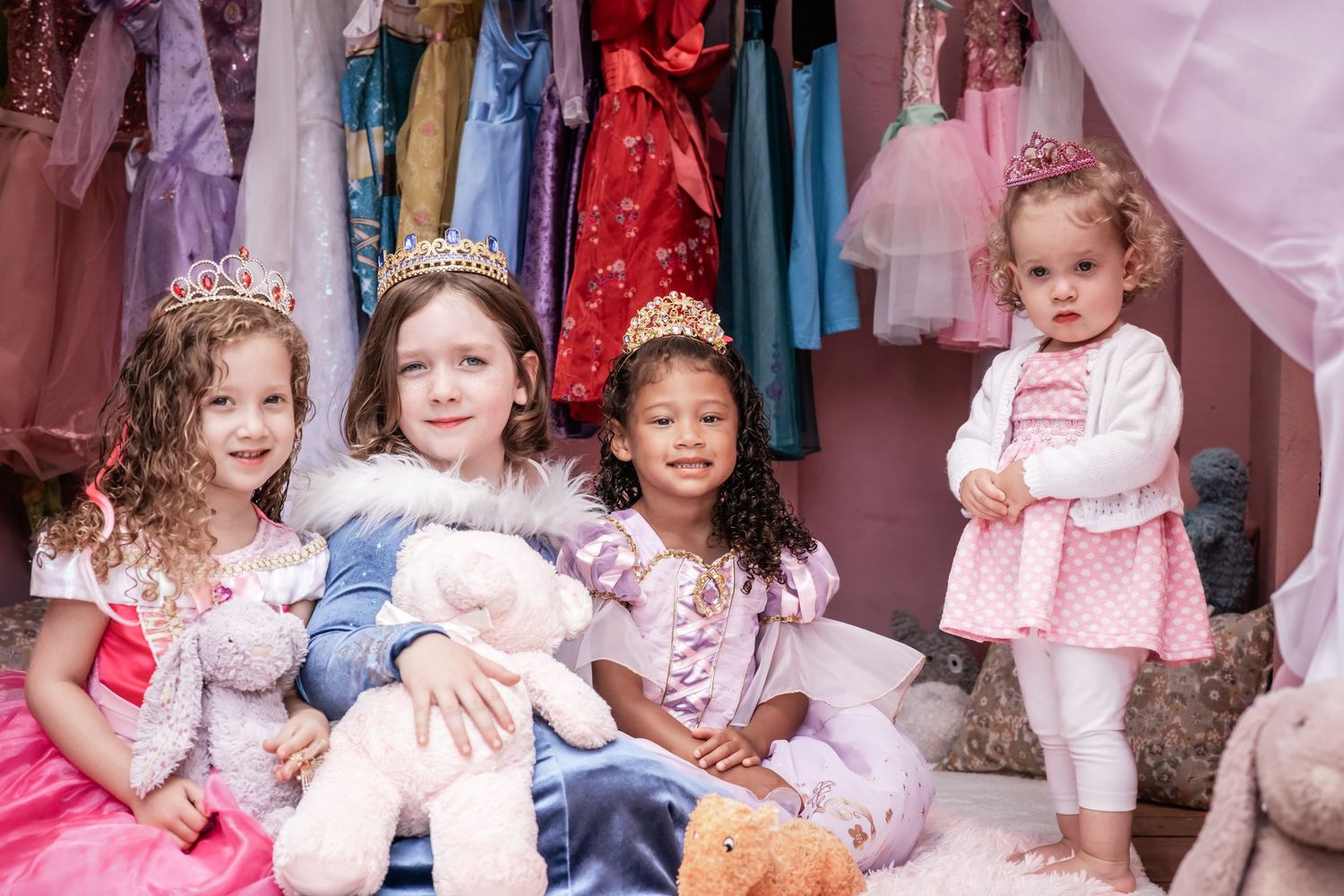 Four young children wearing fancy dress costumes and princess dresses sit together against a backdrop of colorful clothing.
