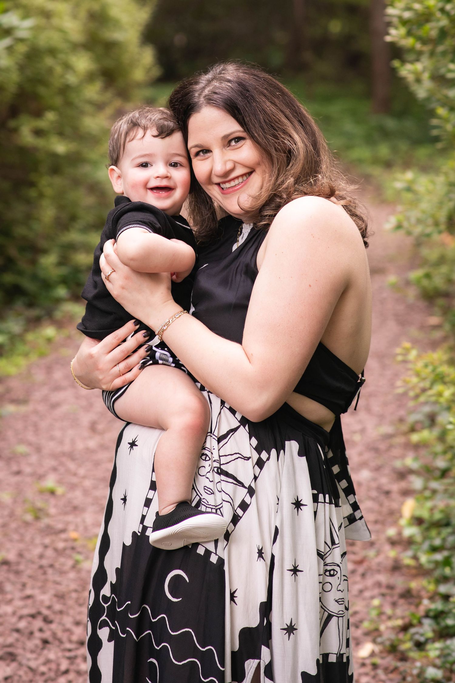 A smiling woman in a black and white dress holds a happy baby boy outdoors on a wooded path.
