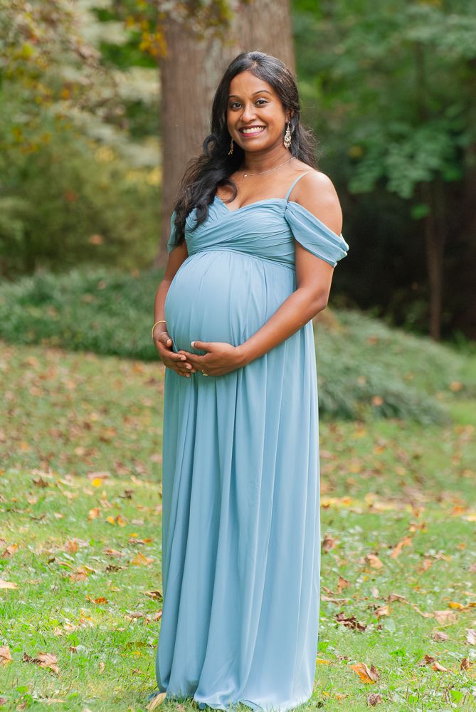 A pregnant woman in a light blue off-shoulder maxi dress poses outdoors on grass with autumn leaves in the background.