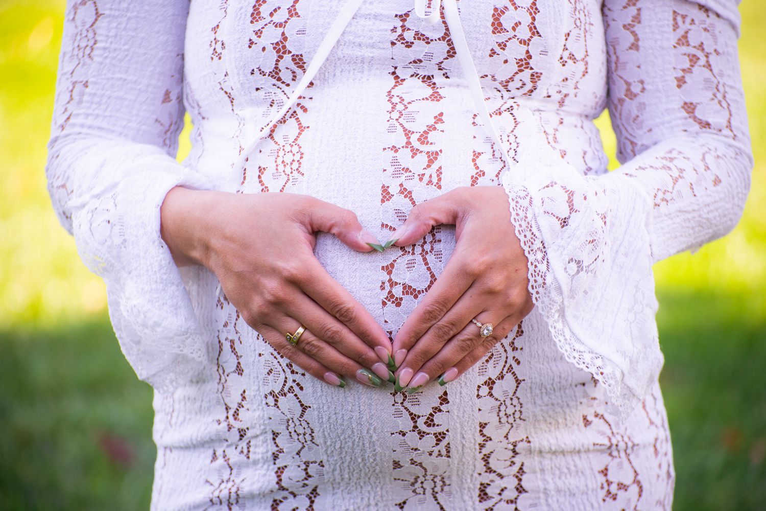 Pregnant woman in white lace dress forming heart shape with hands on baby bump outdoors.