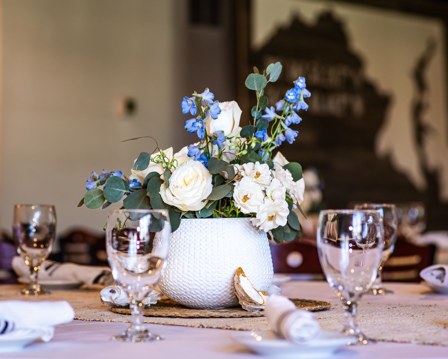 White vase with roses and blue flowers as elegant wedding table centerpiece with crystal glasses.