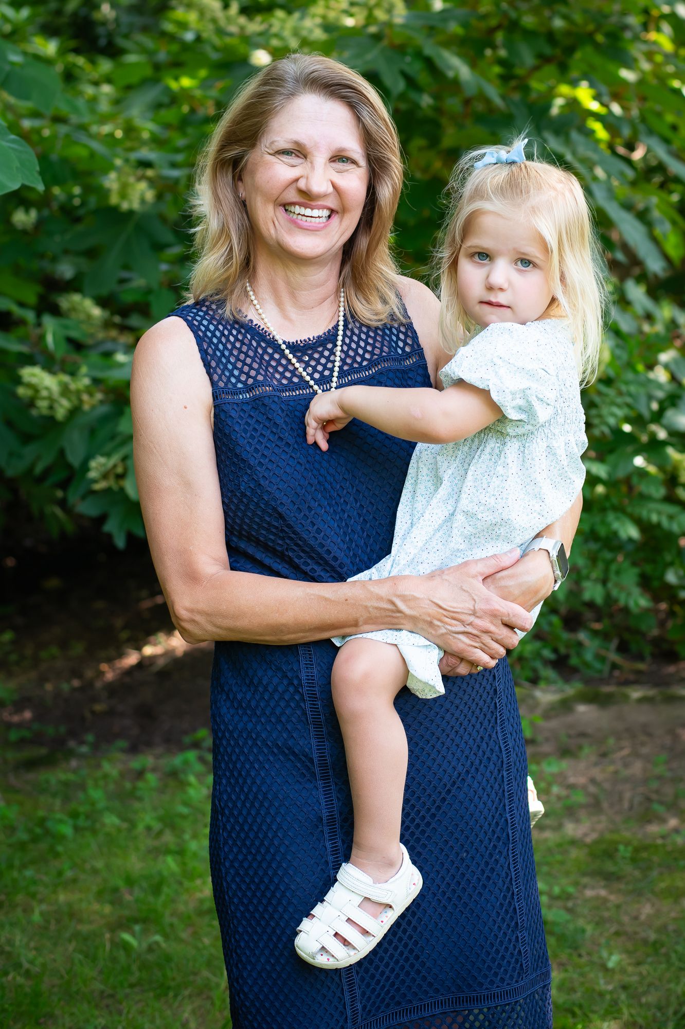 Smiling woman in navy dress holding a young blonde girl in white dress outdoors in a green garden setting.