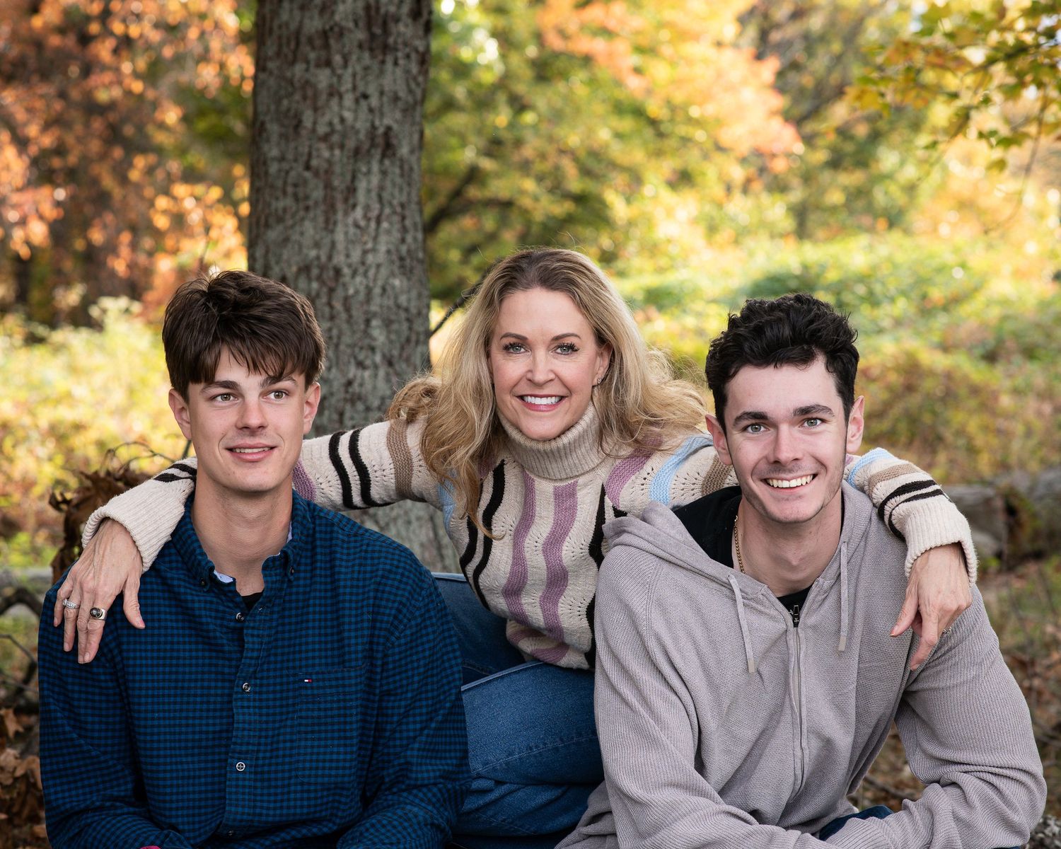 A smiling woman poses with two young men outdoors amid colorful autumn foliage for a fall family portrait.