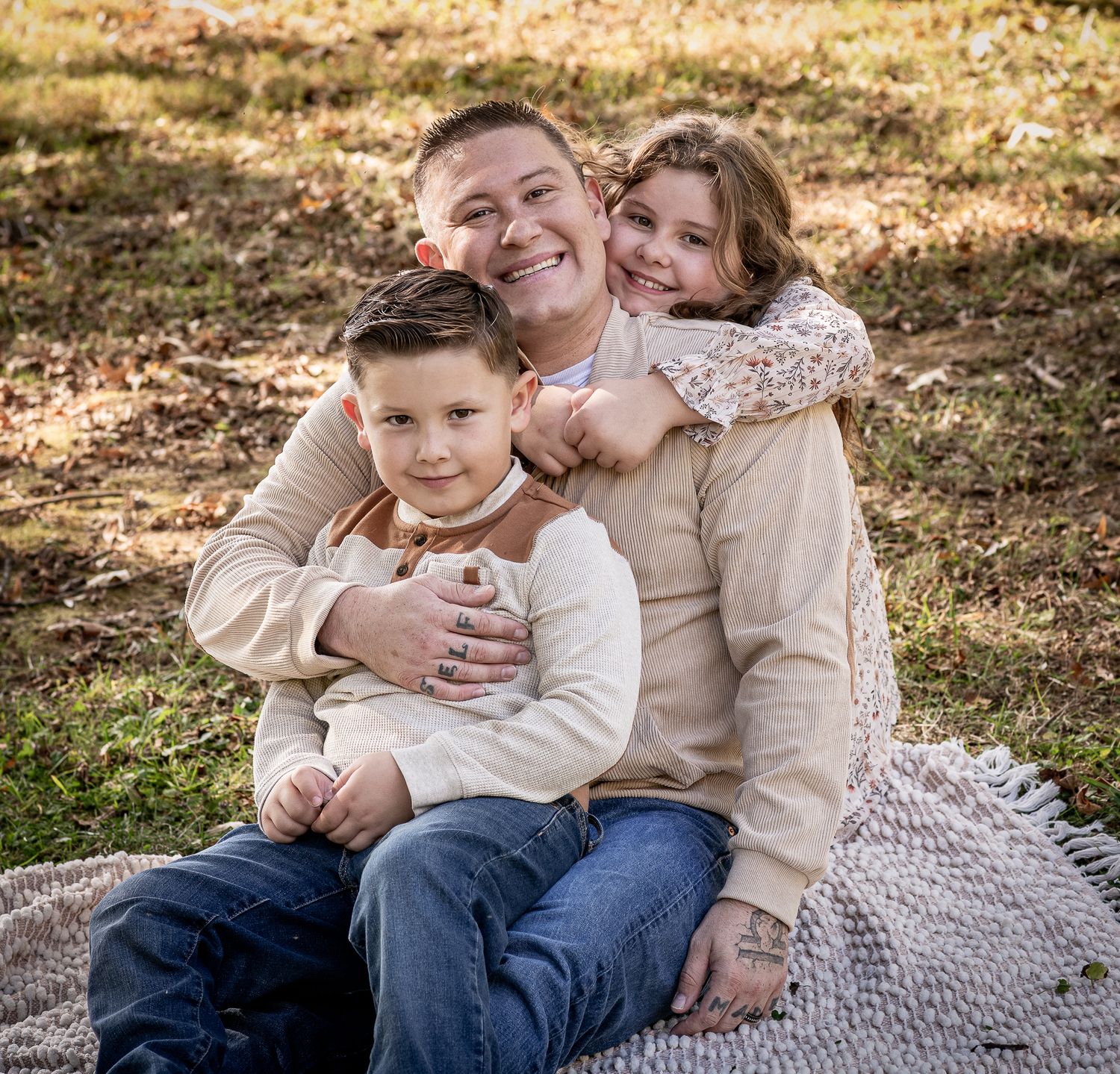 A smiling man sits outdoors on a blanket with two young children hugging him in a warm fall family portrait.