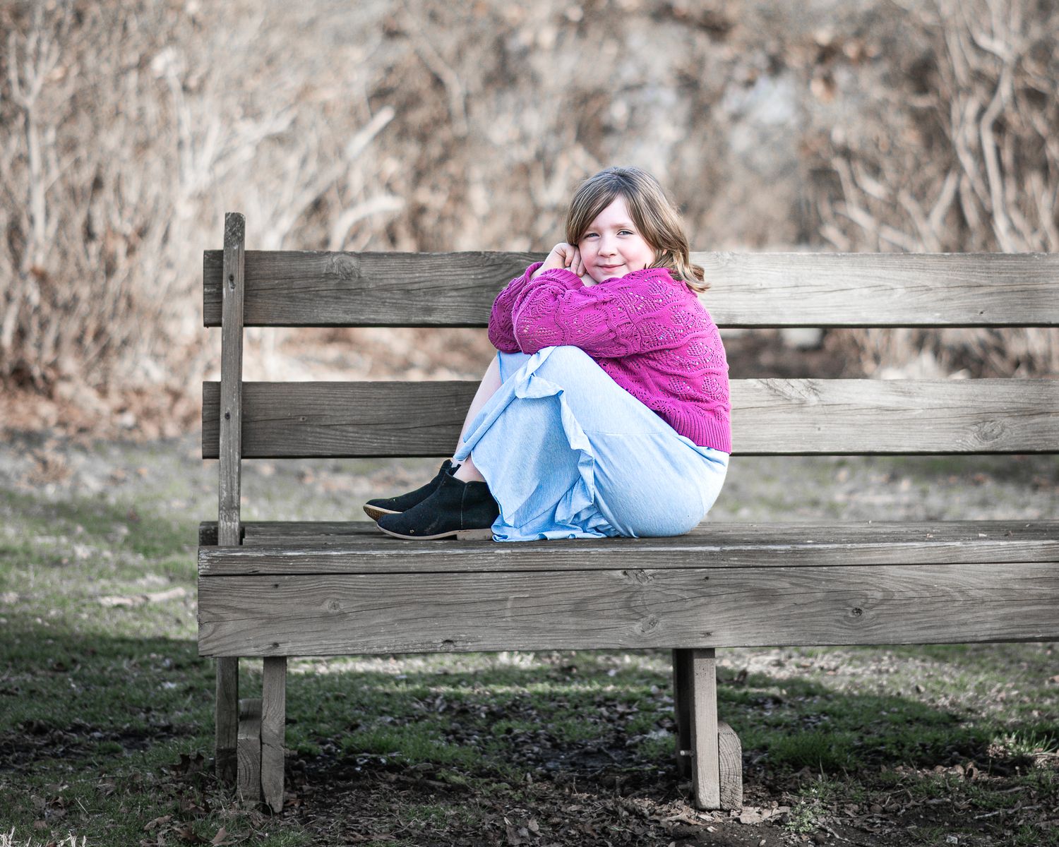 A person in a pink sweater and jeans sits curled up on a wooden park bench on a winter day.